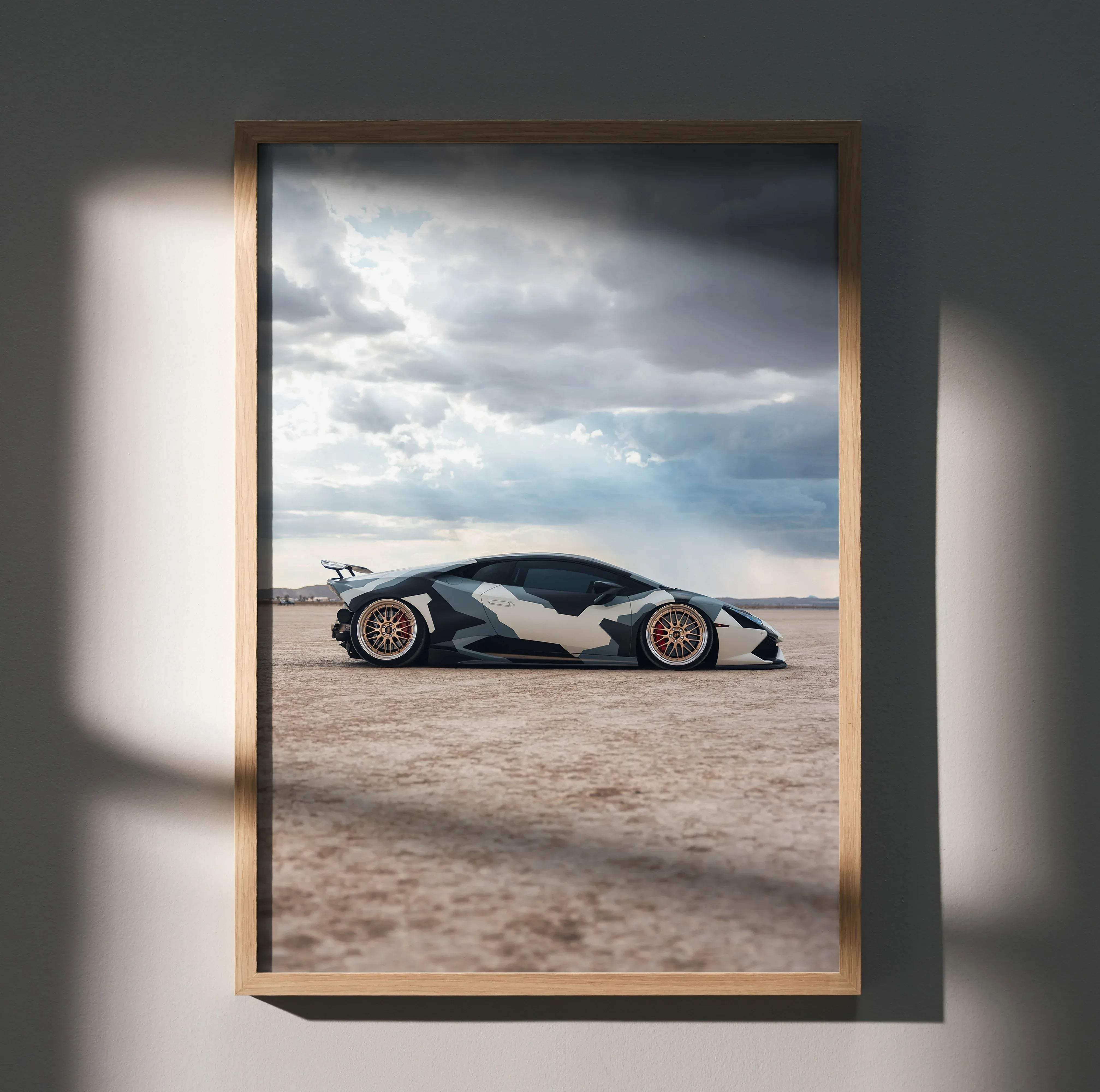 A framed poster of a Lamborghini Huracan with black and white camouflage against a cloudy desert sky