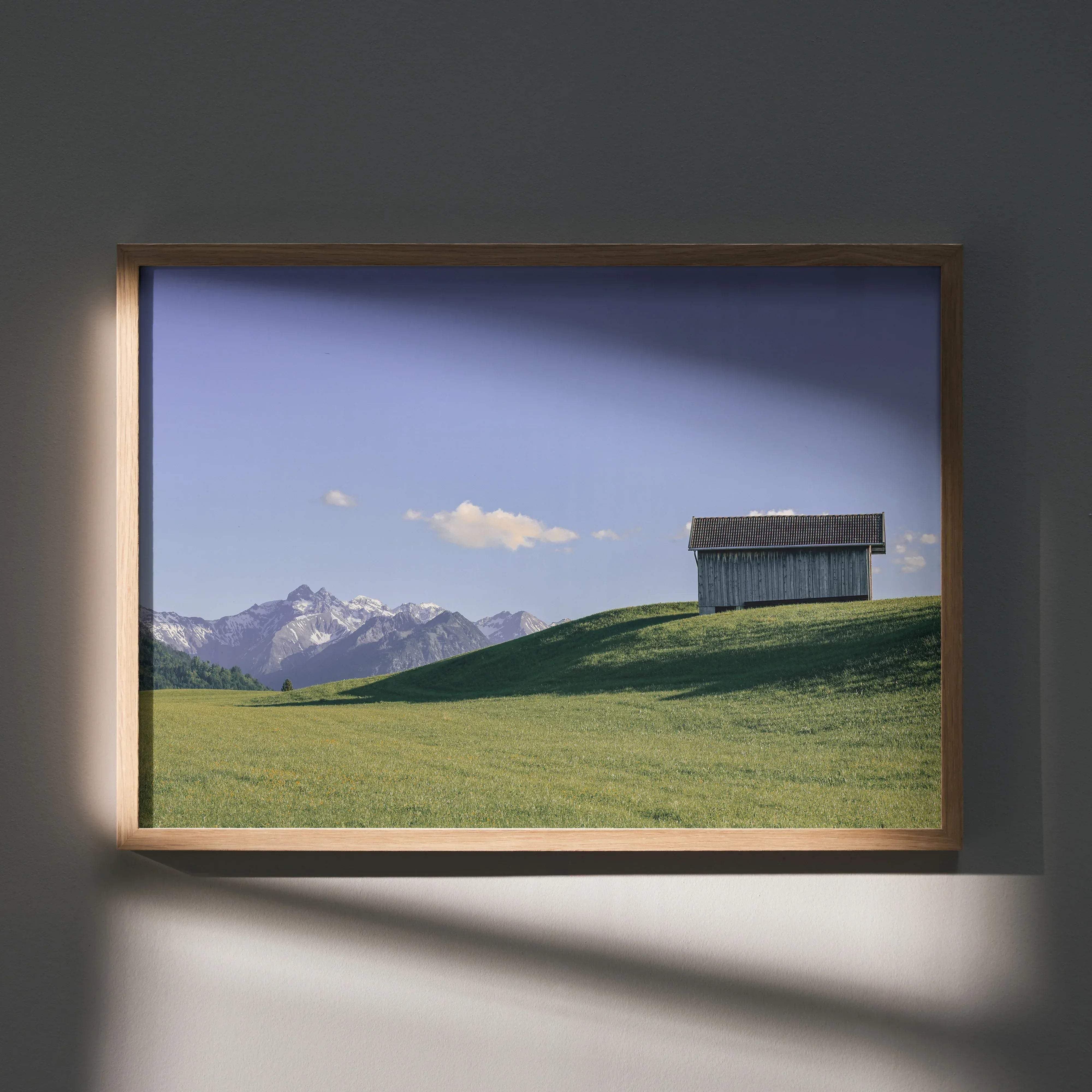 A framed landscape print of a rustic wooden shed on a green hill with snow-capped mountains in a light wood frame