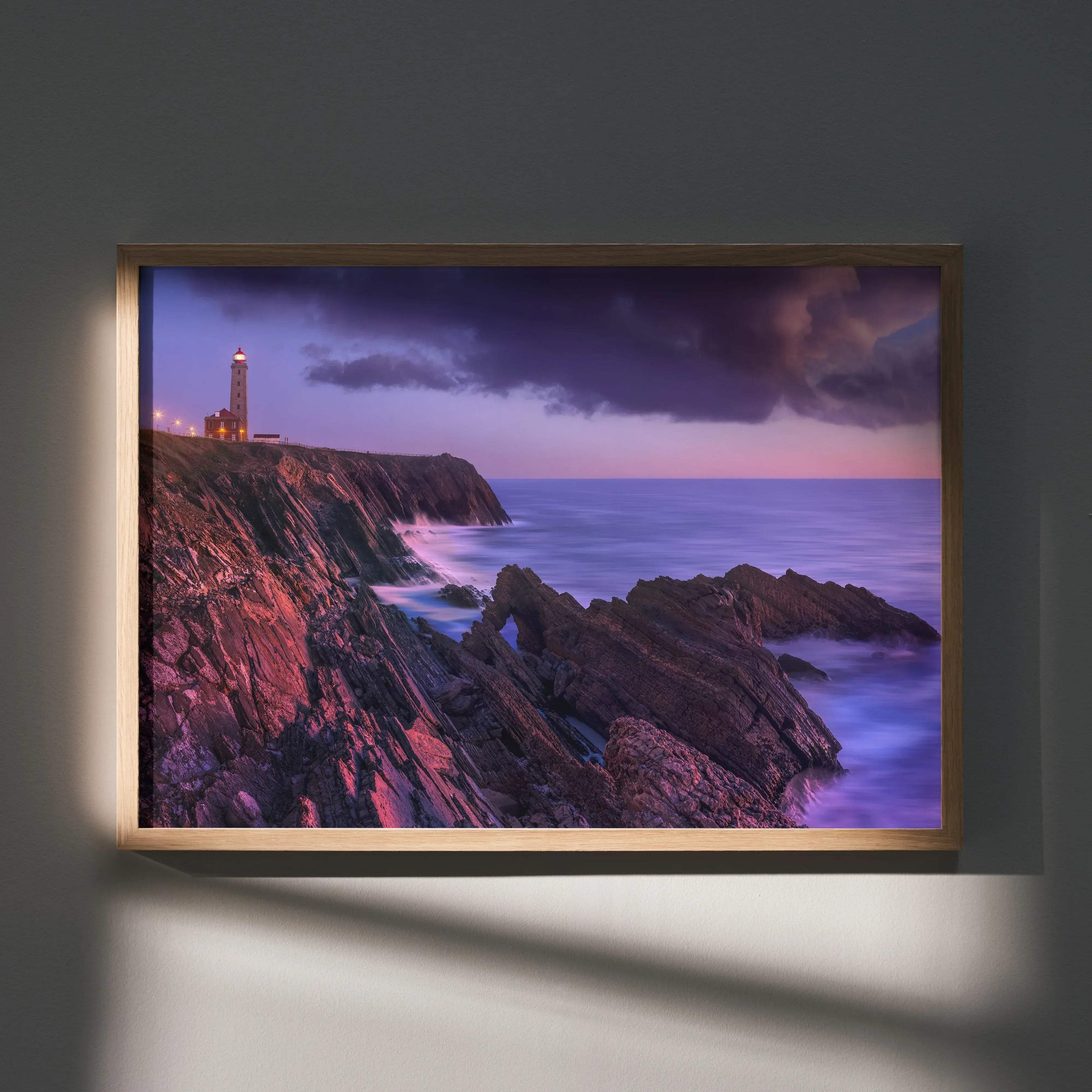A framed coastal landscape art poster featuring a lighthouse on cliffs under a purple and pink twilight sky in a light wooden frame