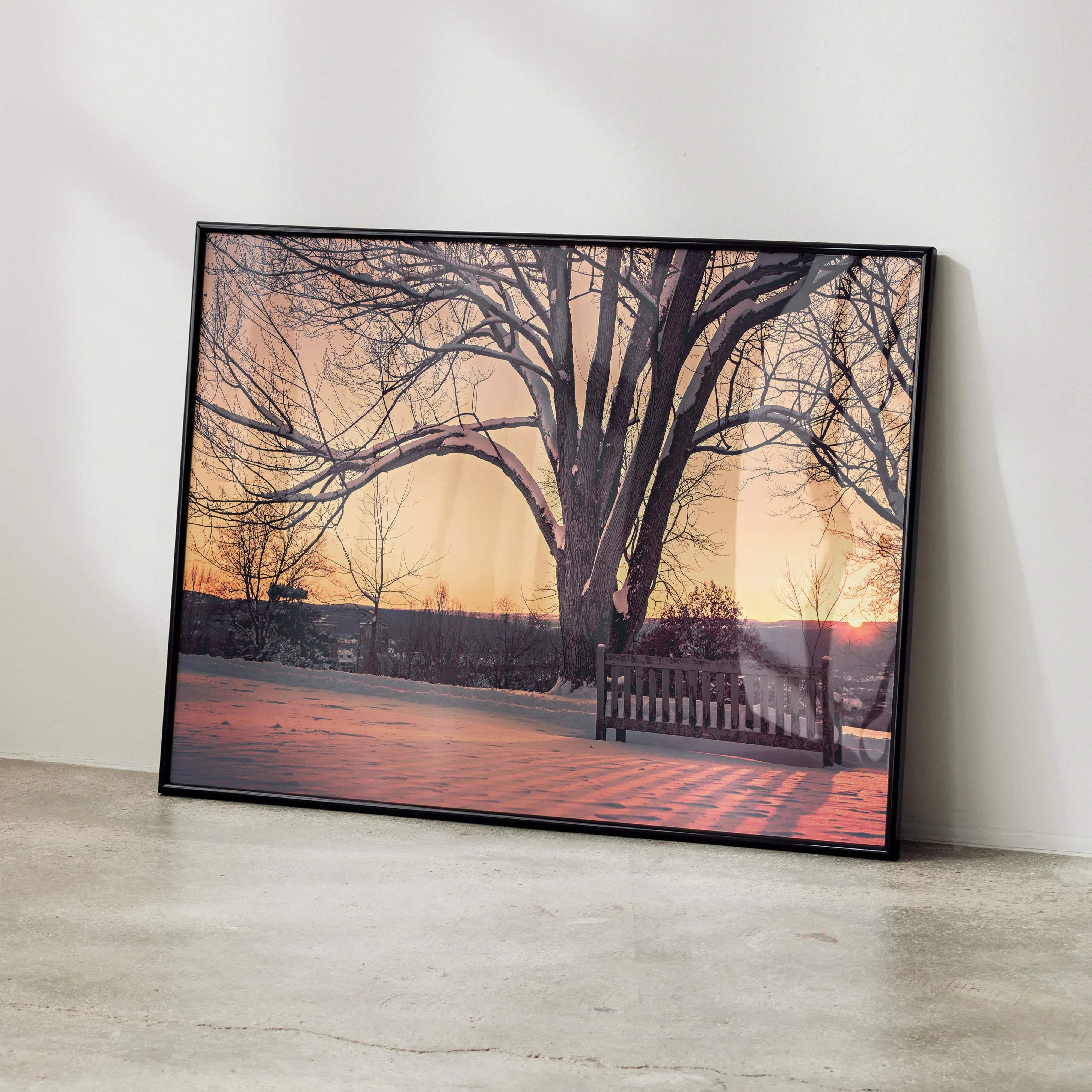 Black-framed canvas print of serene snowy landscape with snow-dusted tree and bench at warm sunset hues
