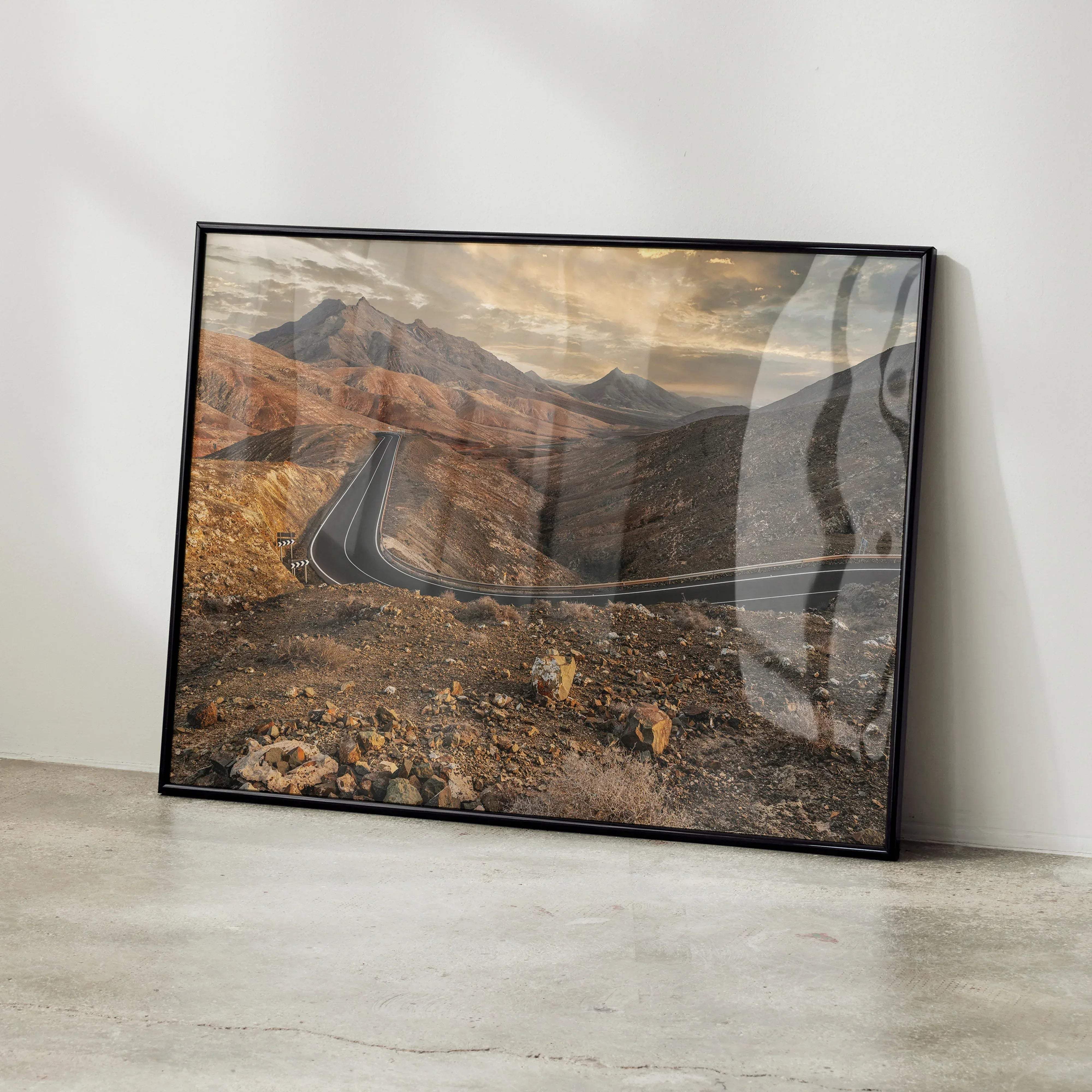 Black-framed landscape print of a winding road through sunlit rugged mountains with earthy tones for wall art decor