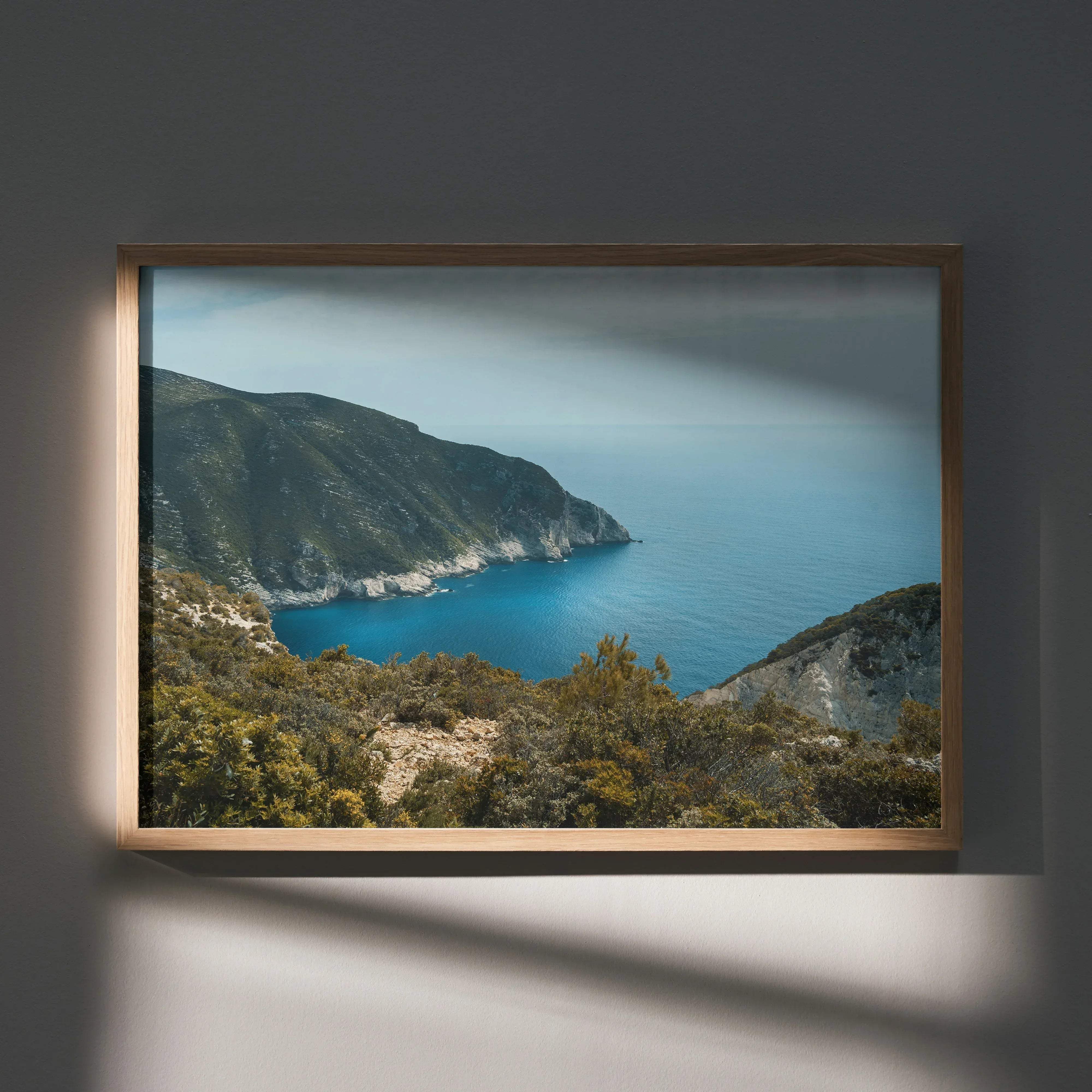 A rectangular framed wall art poster with a light wood frame featuring a coastal landscape of deep blue water and green hills