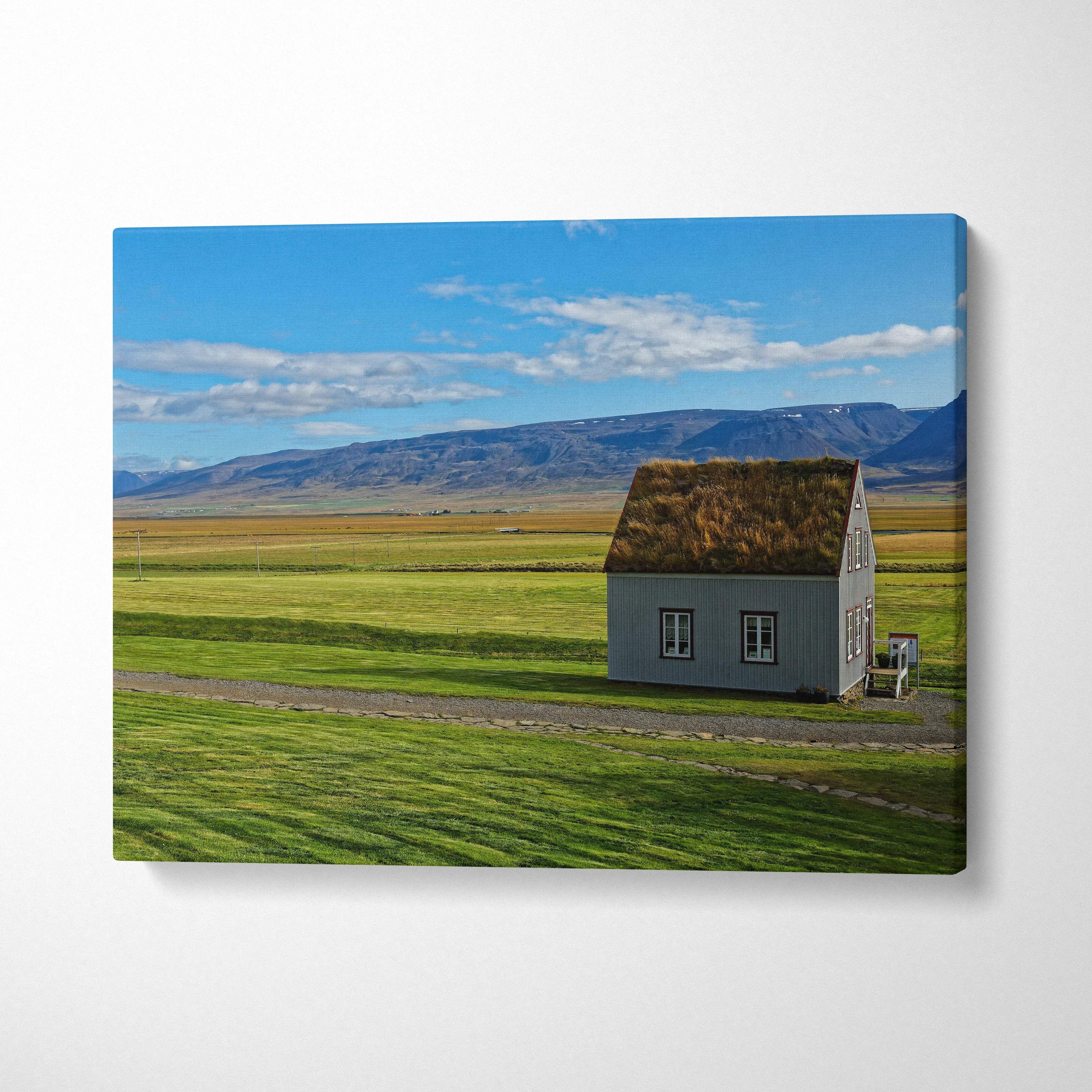 Canvas wall art featuring a solitary house in a green field under a bright blue sky with mountains in the background.