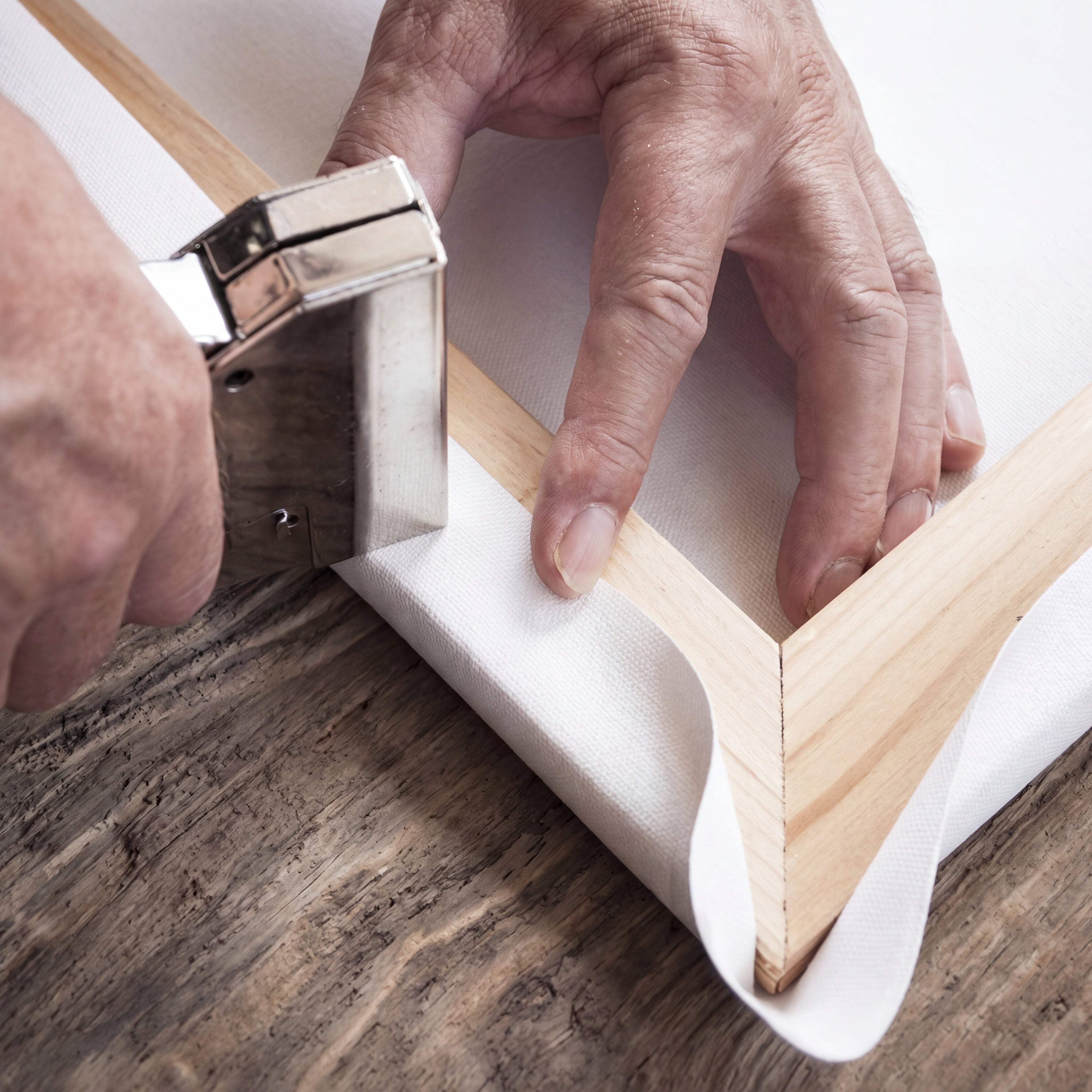 Silver metal canvas stretcher bar being used to secure a white fabric canvas onto a light wood frame.