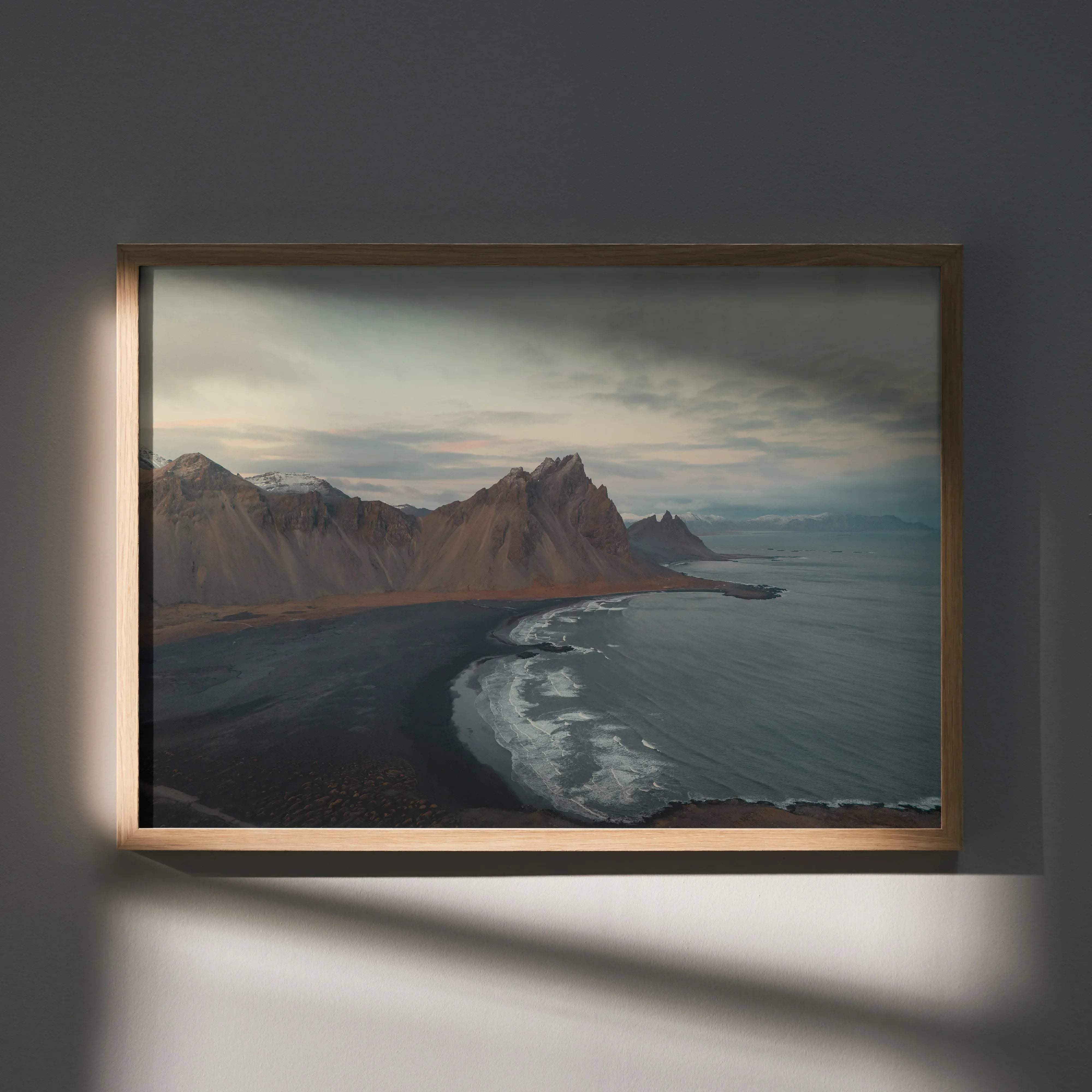 A framed landscape photograph print of a rugged coastal scene with snow-capped mountains and dark waves in a light wood frame