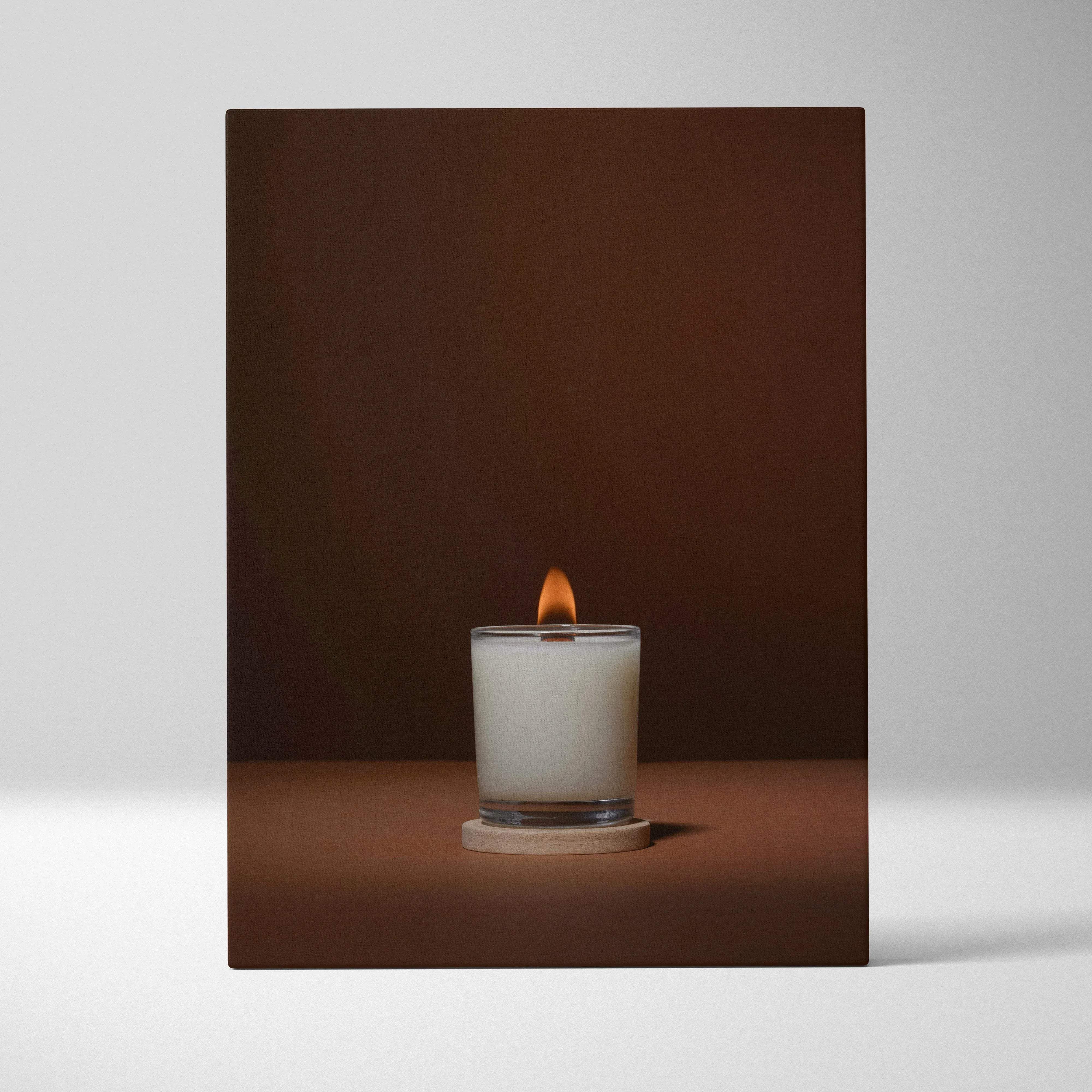 Lit white scented candle in clear glass jar on wooden coaster with warm glow
