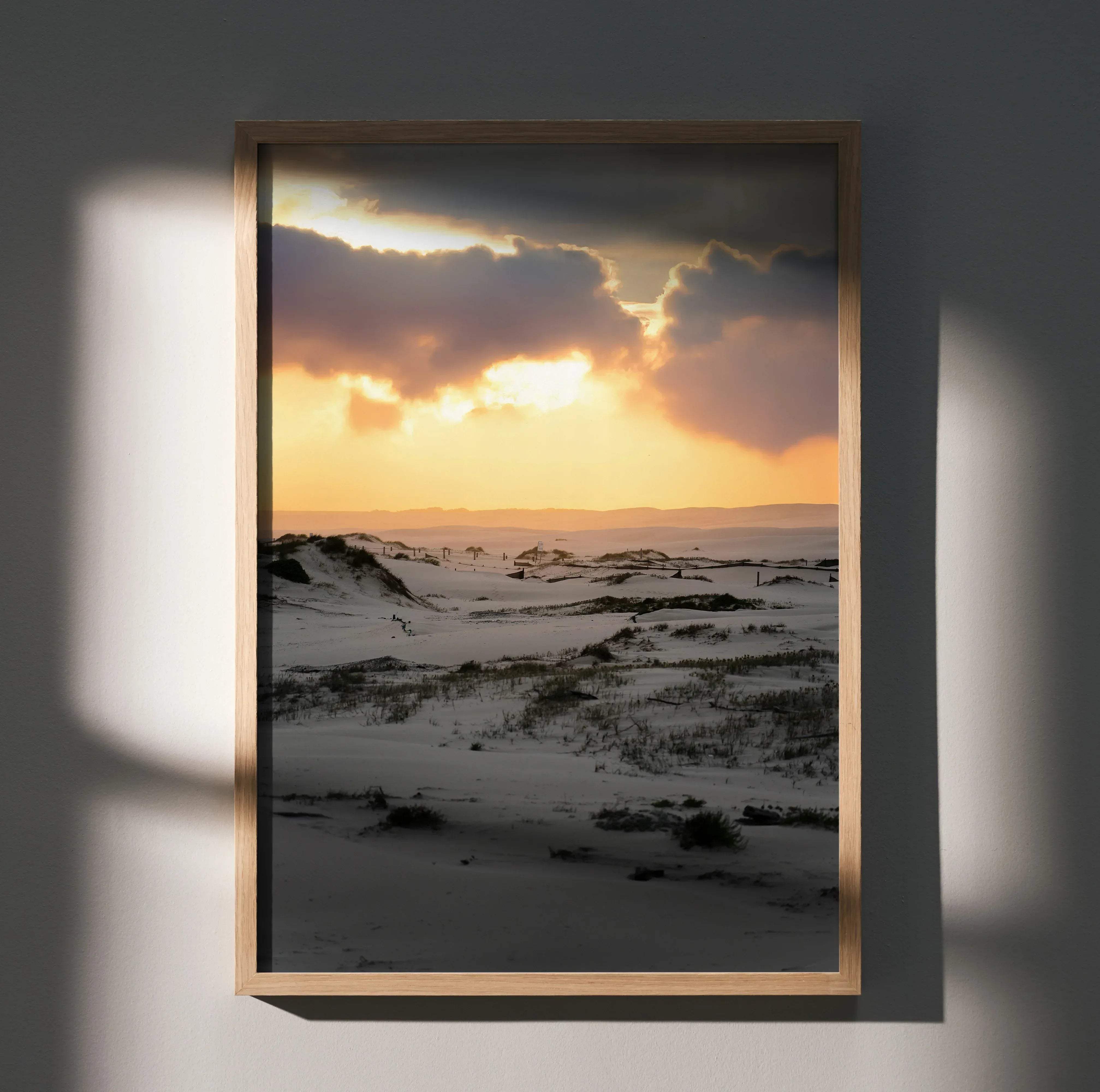 A rectangular wooden-framed art print showing a dramatic sunset over snow-covered dunes with dark clouds
