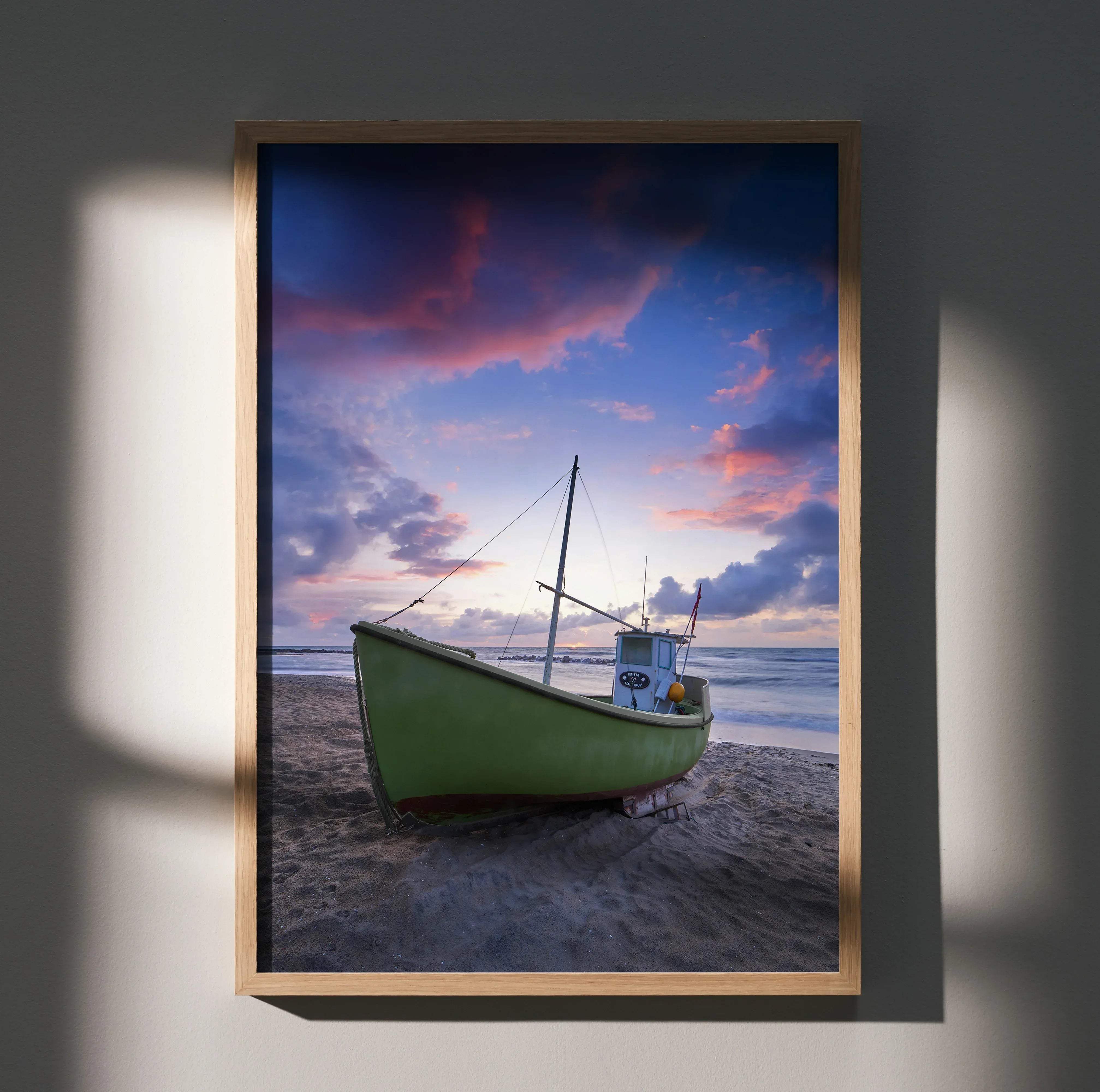 A framed vertical art print poster of a green fishing boat on a sandy beach under pink and purple sunset clouds