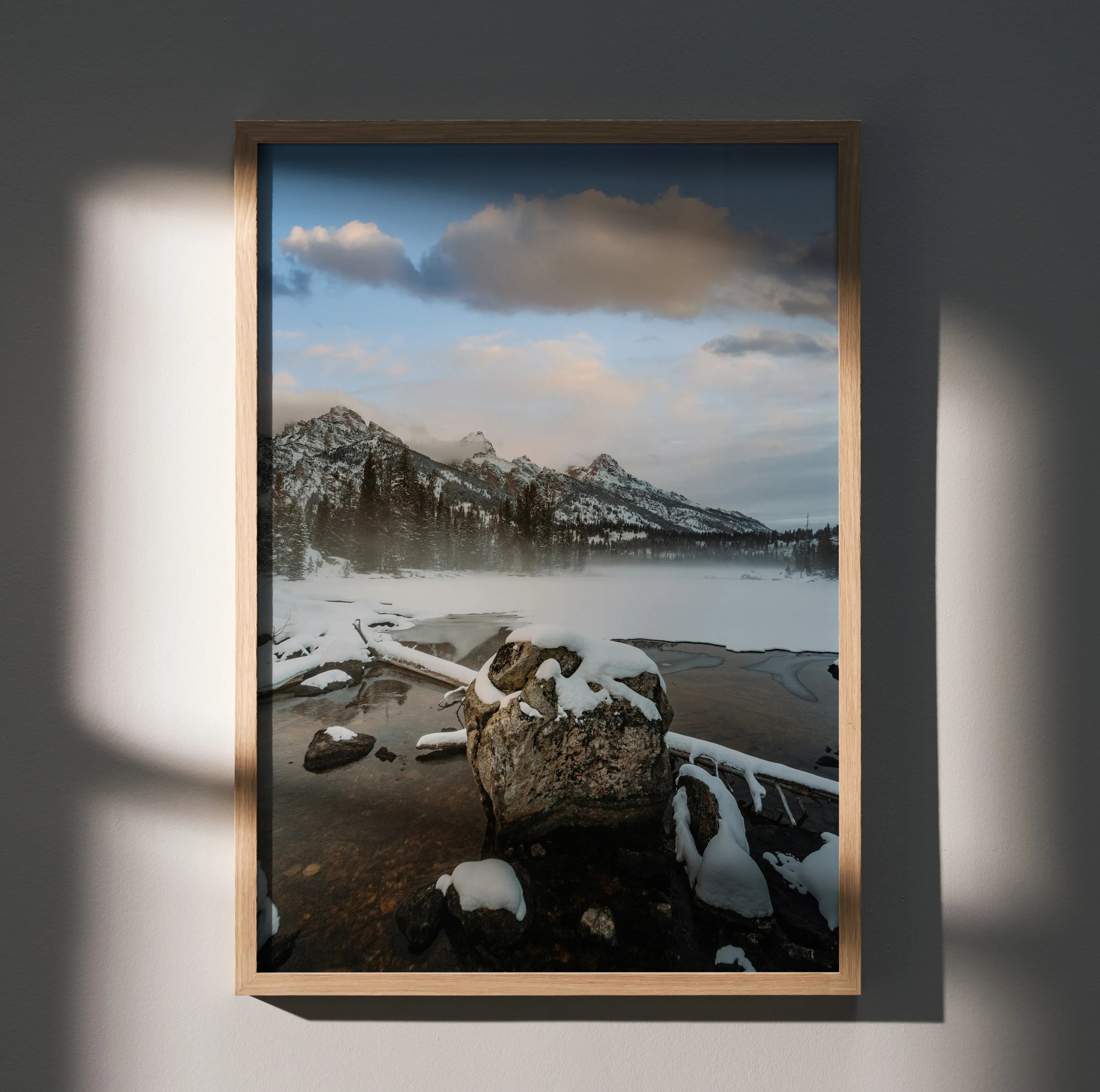 Vertical poster wall art featuring snowy mountain peaks under a cloudy sky over a calm lake with rocks and driftwood.