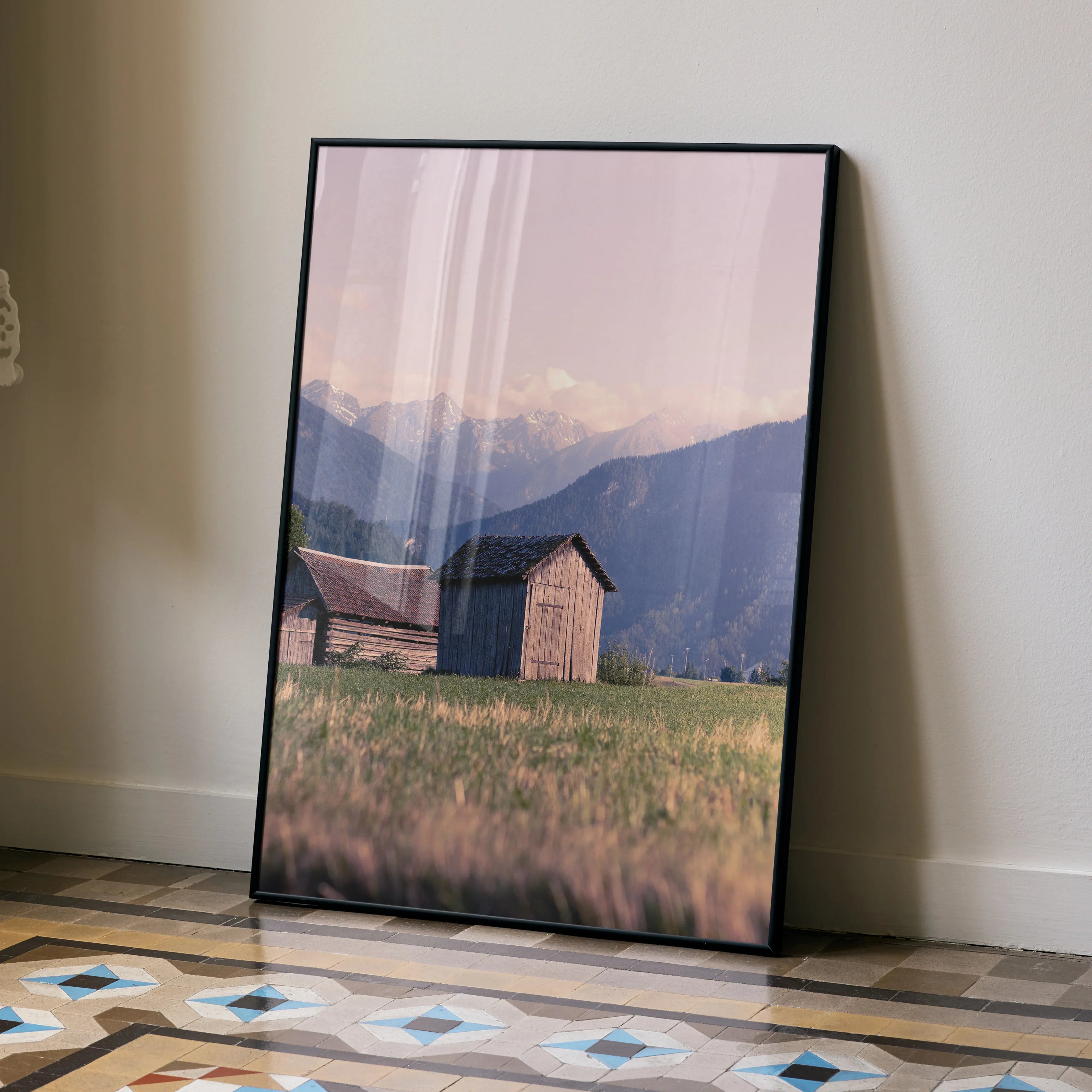 Vertical wall art poster featuring rustic wooden cabins in a grassy field with layered blue mountains in the background.