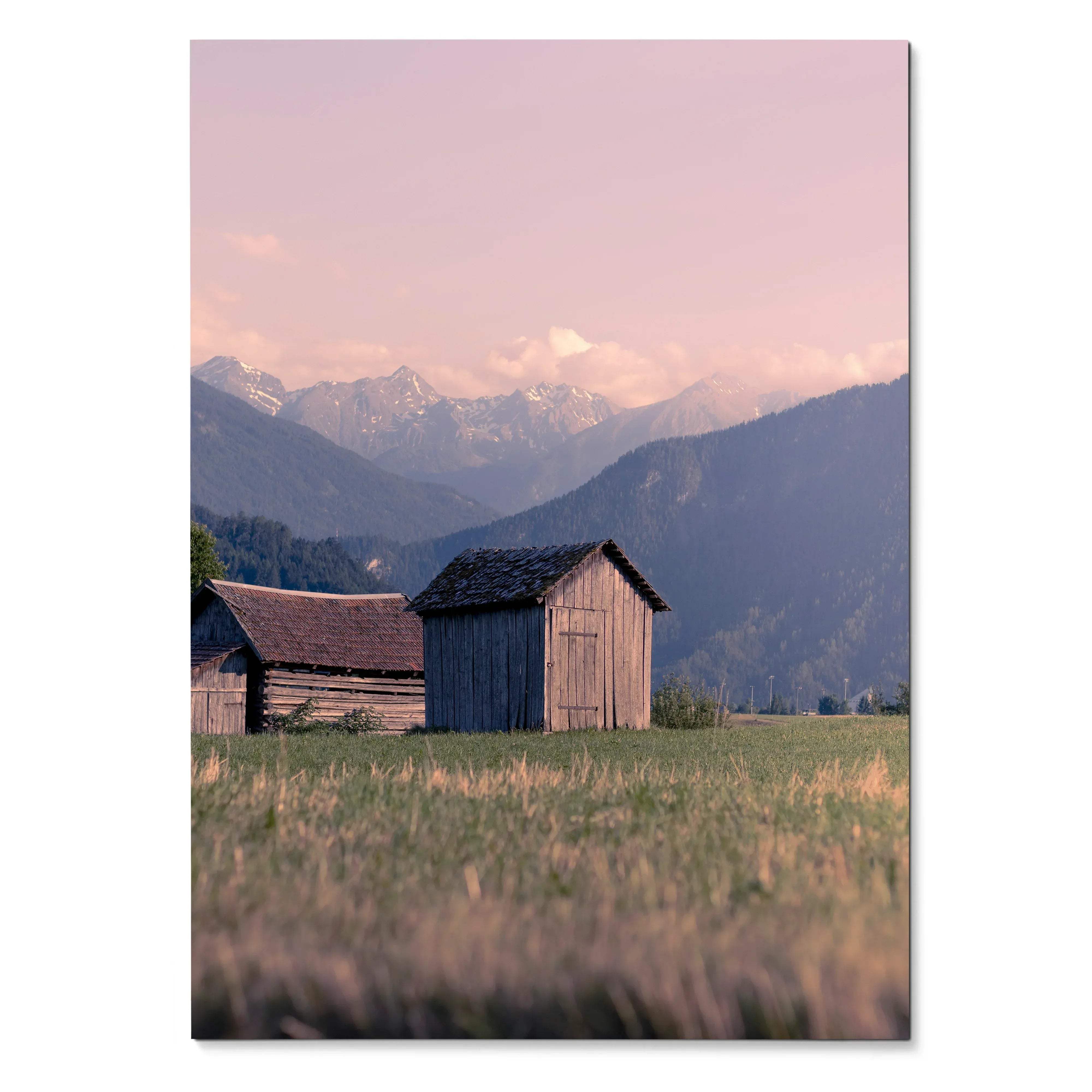 Vertical poster wall art of rustic wooden barns in a grassy field with mountain range and soft pink sky background.
