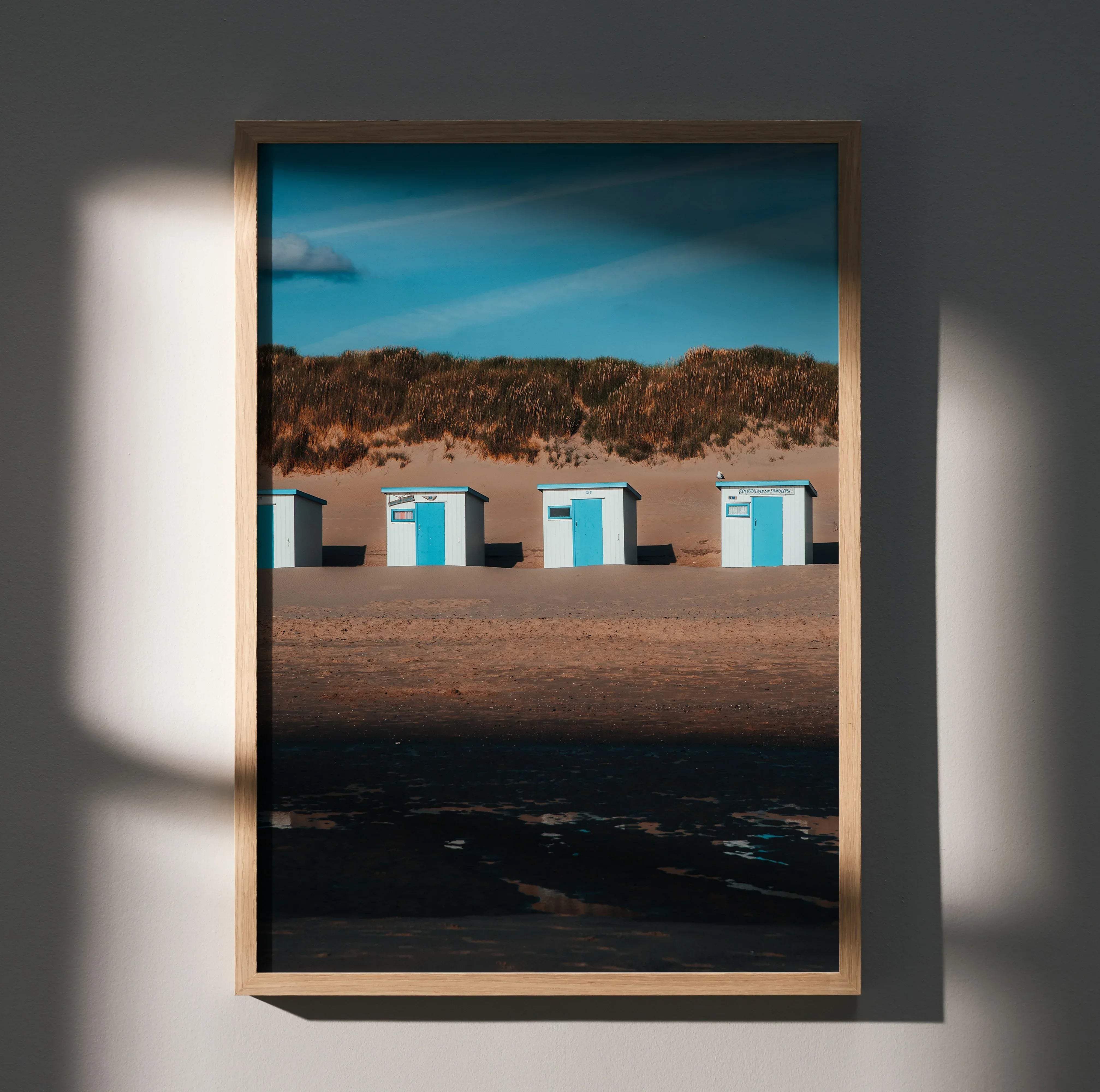 A framed serene beach landscape wall art poster featuring four white beach huts with bright blue doors on sand dunes