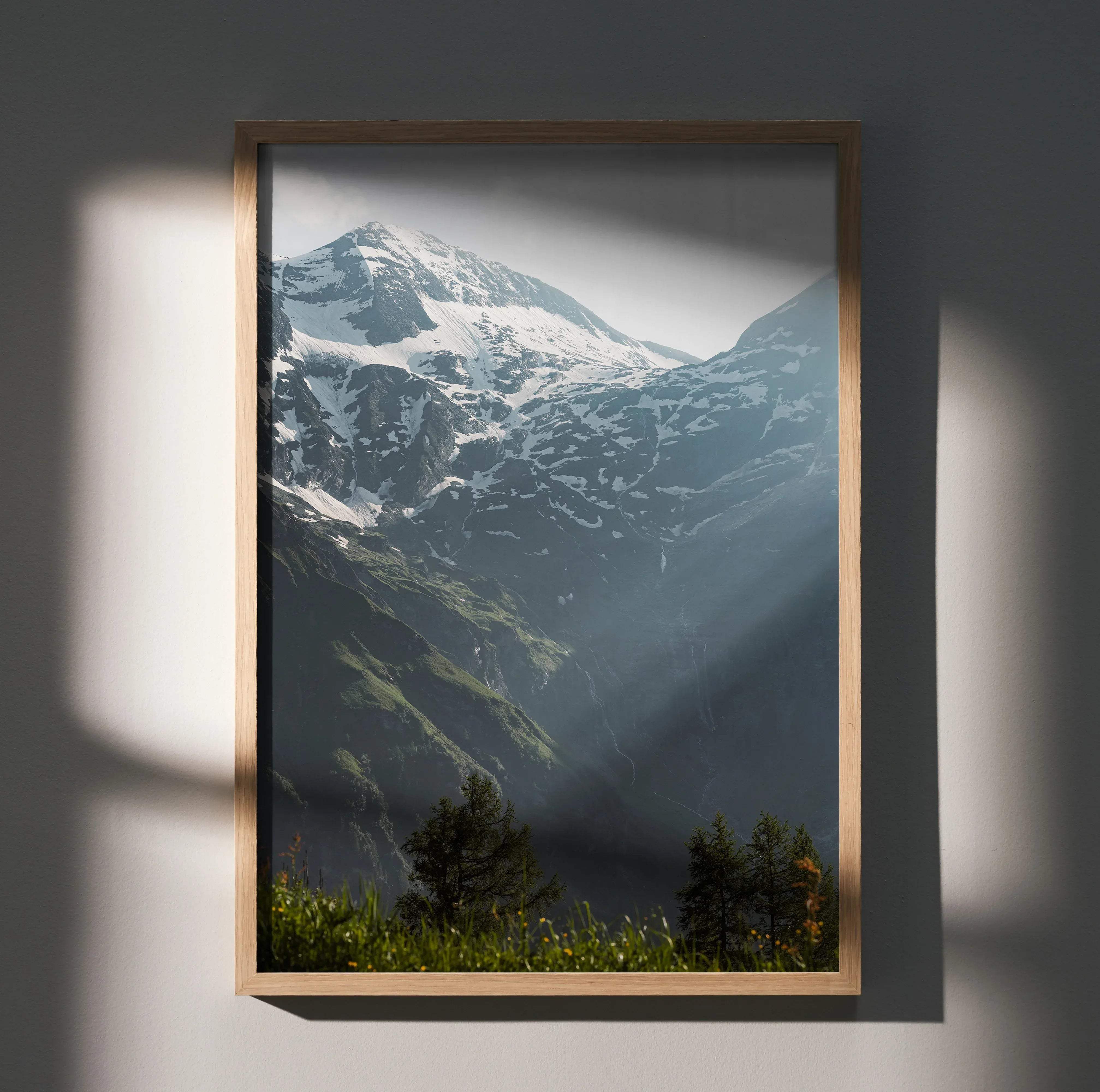 A framed vertical mountain landscape print with a light wood frame showing snow-capped peaks and green valleys