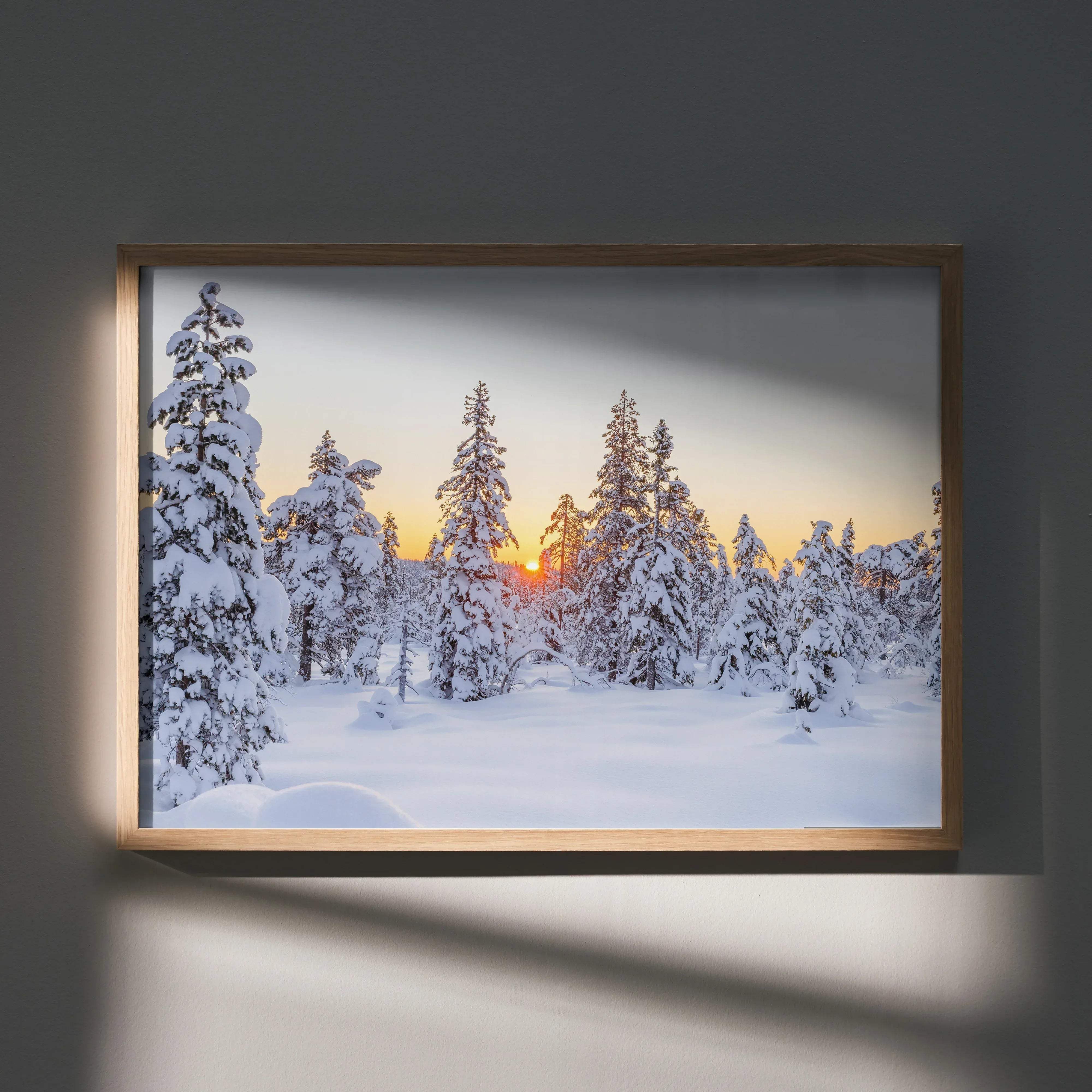 A framed winter landscape art poster featuring snow-covered pine trees against a soft orange and gray sunset sky in a light wood frame