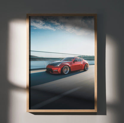 Red Nissan 370Z driving on coastal road wall art poster in wooden frame.