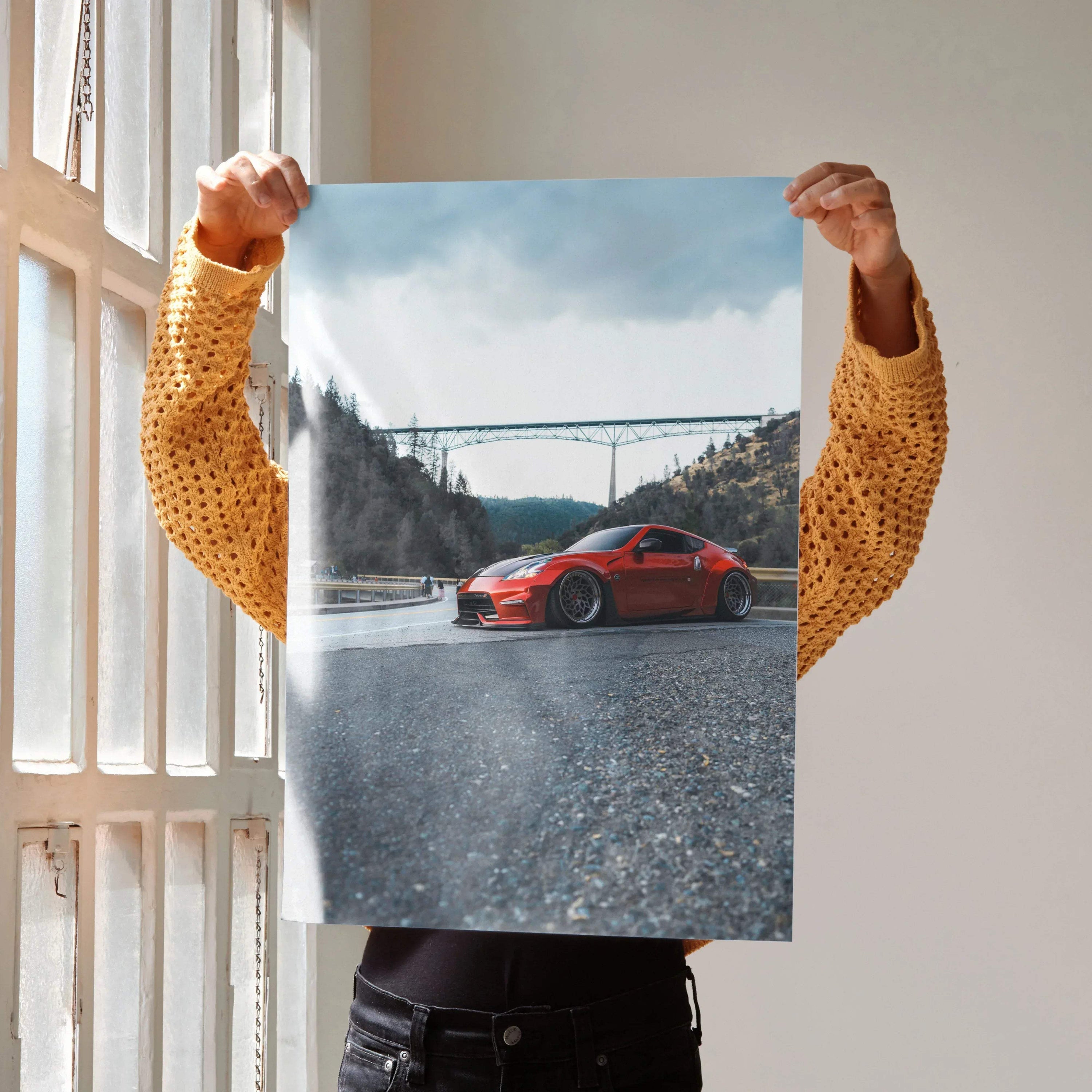 Red Nissan 370Z poster on textured road with bridge and cloudy sky background wall art.