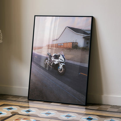 BMW S1000RR motorcycle poster leaning on floor with rustic building and sunset sky, wall art for enthusiasts.