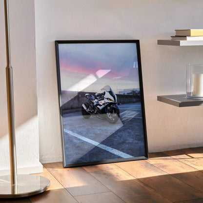 BMW S1000RR Drag Spec motorcycle poster in black frame leaning on floor, wall art with sunset sky background.