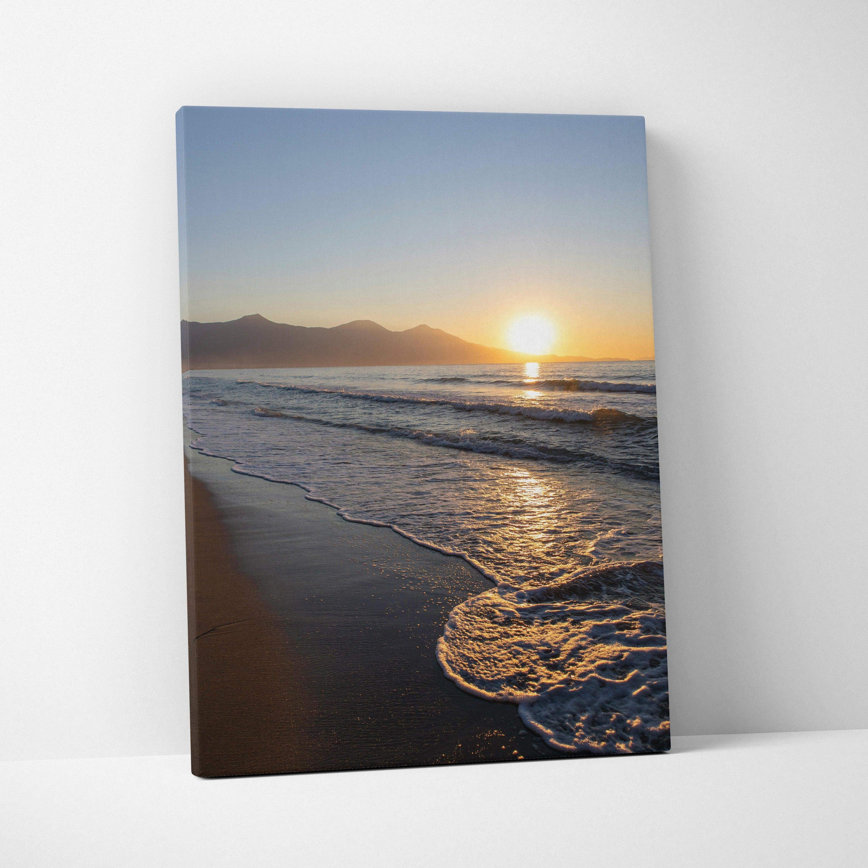 Canvas wall art featuring a sunset over ocean waves with distant mountains and sandy shore.