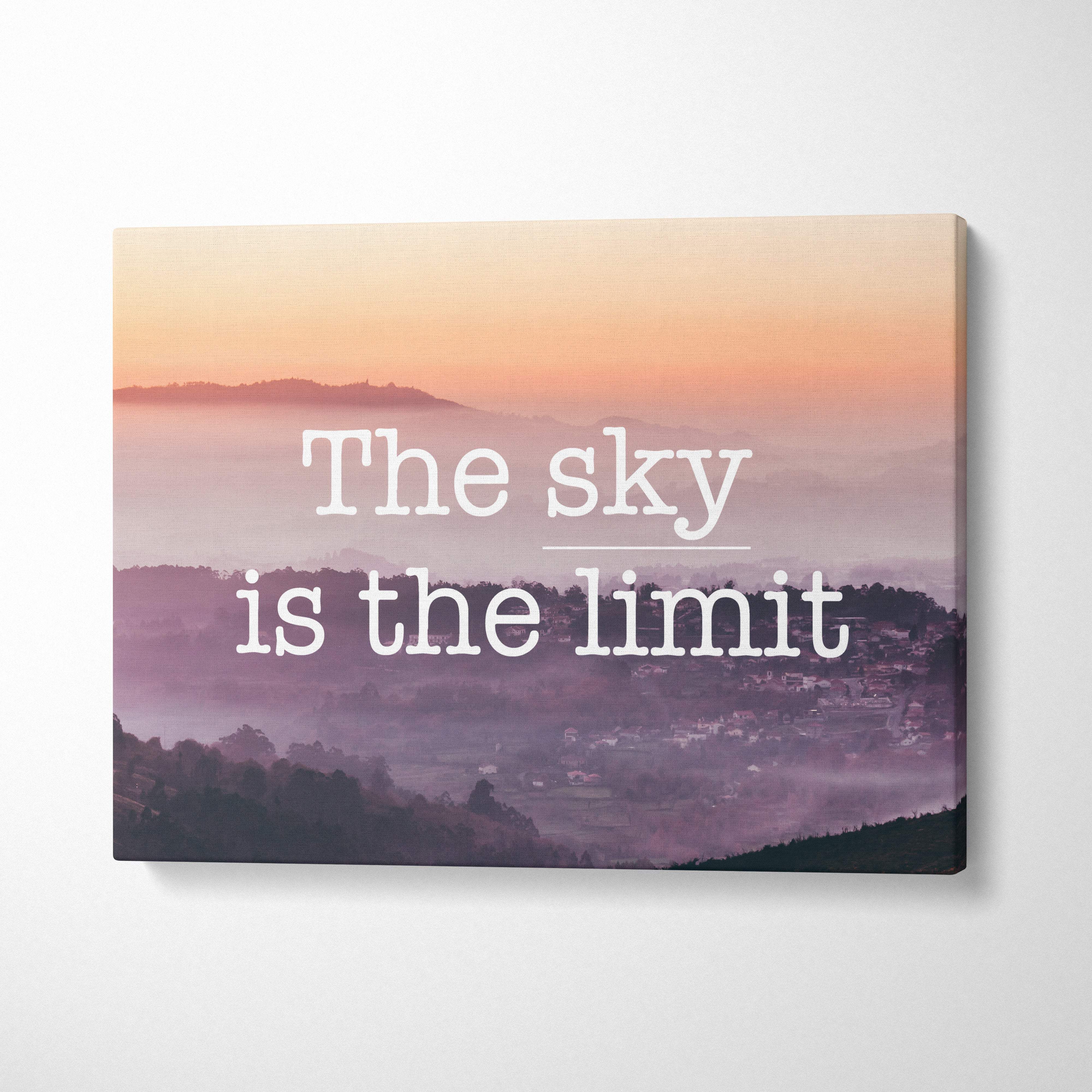 A rectangular canvas print of a misty mountain sunrise with pink and purple hues featuring the sky is the limit text