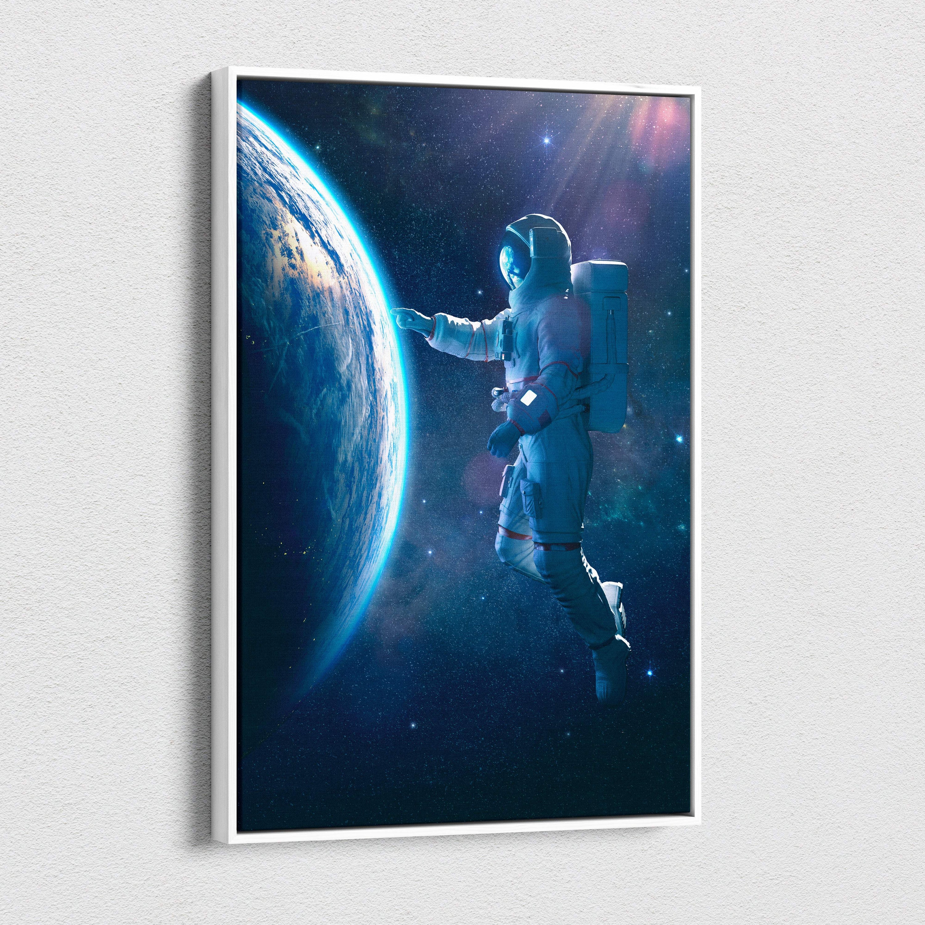A framed blue cosmic space scene canvas featuring a white-suited astronaut reaching toward earth against stars
