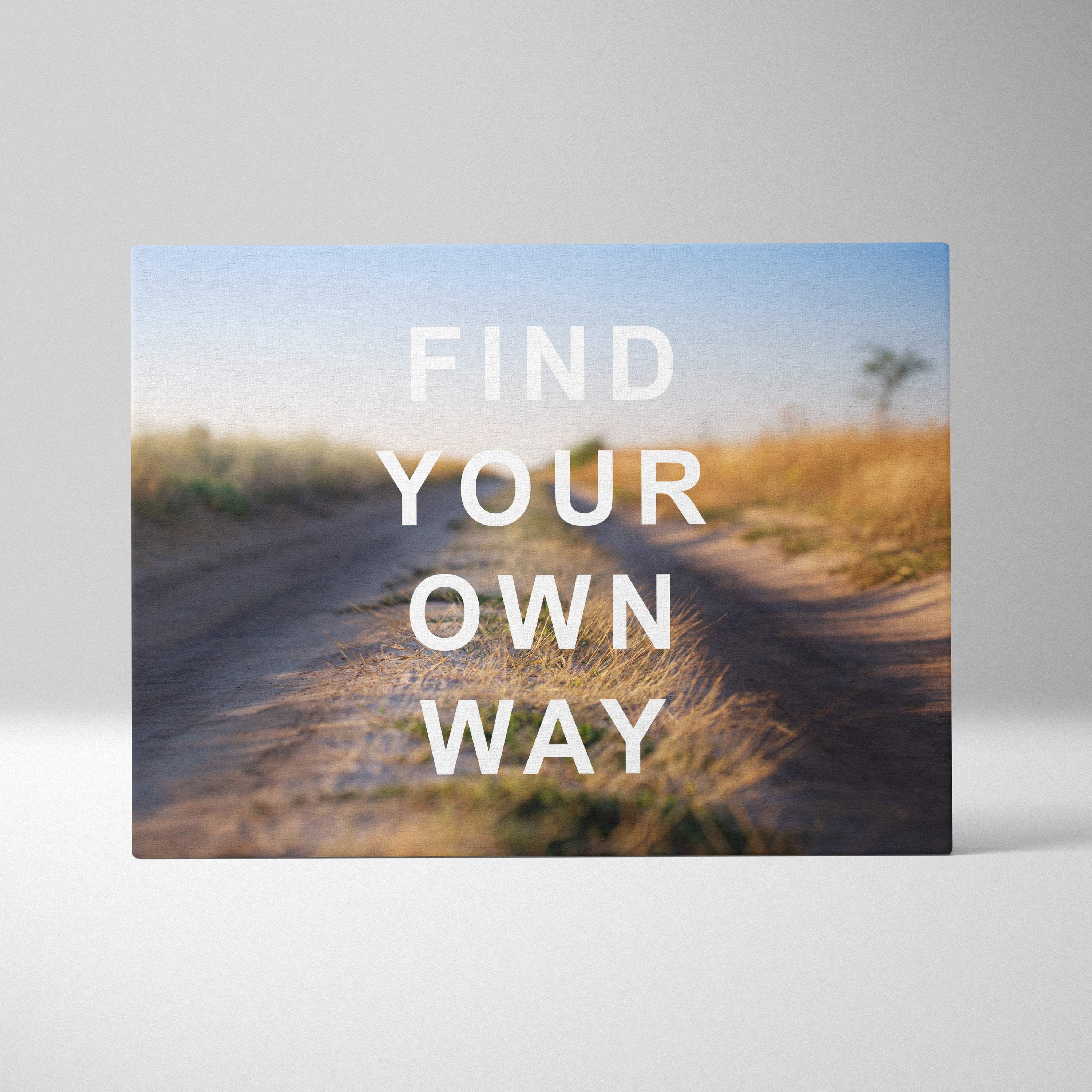 Rectangular canvas print with blurred dirt road through golden grass under blue sky featuring find your own way text