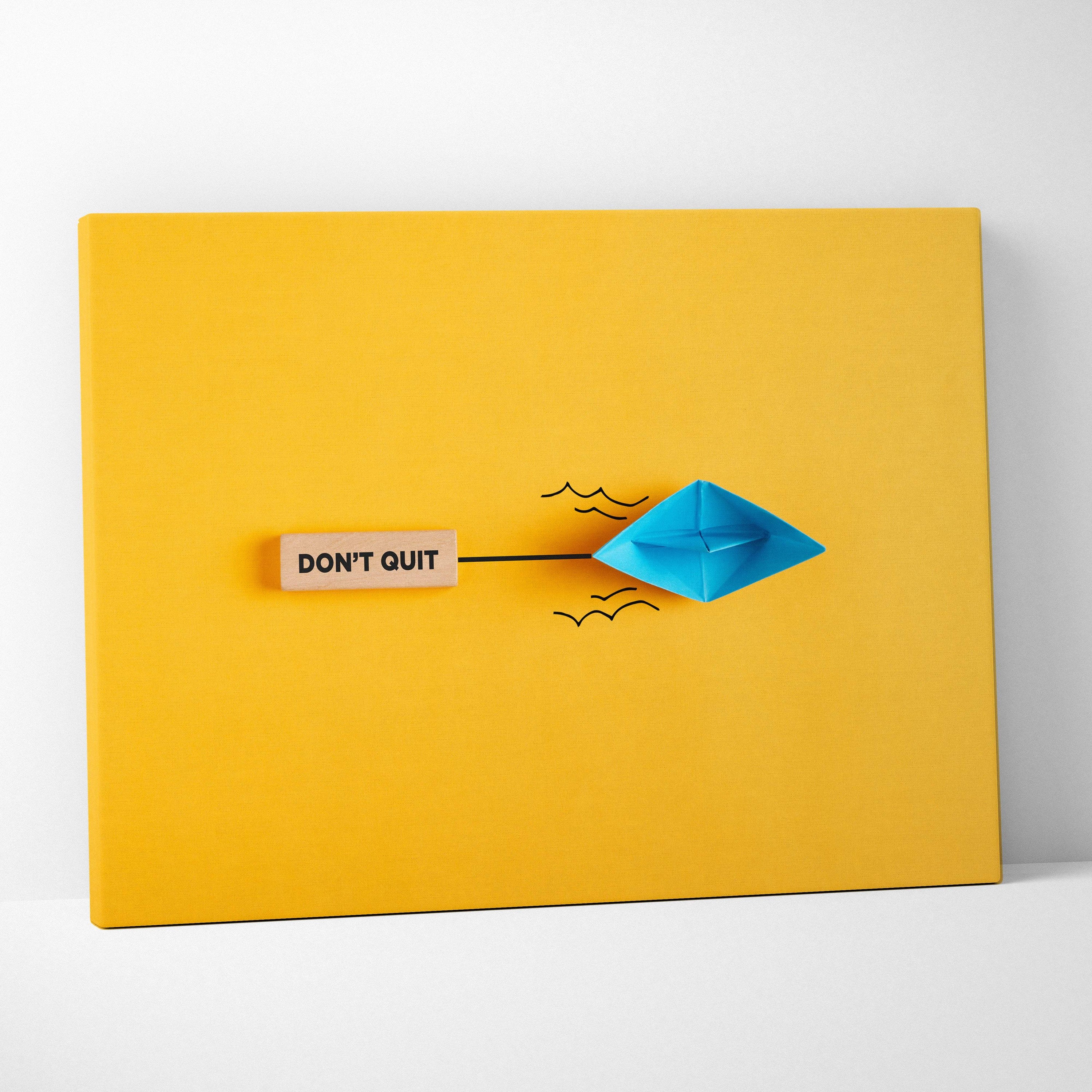 Motivational don’t quit canvas wall art with a blue paper boat black ink waves and wooden plaque on vibrant yellow background