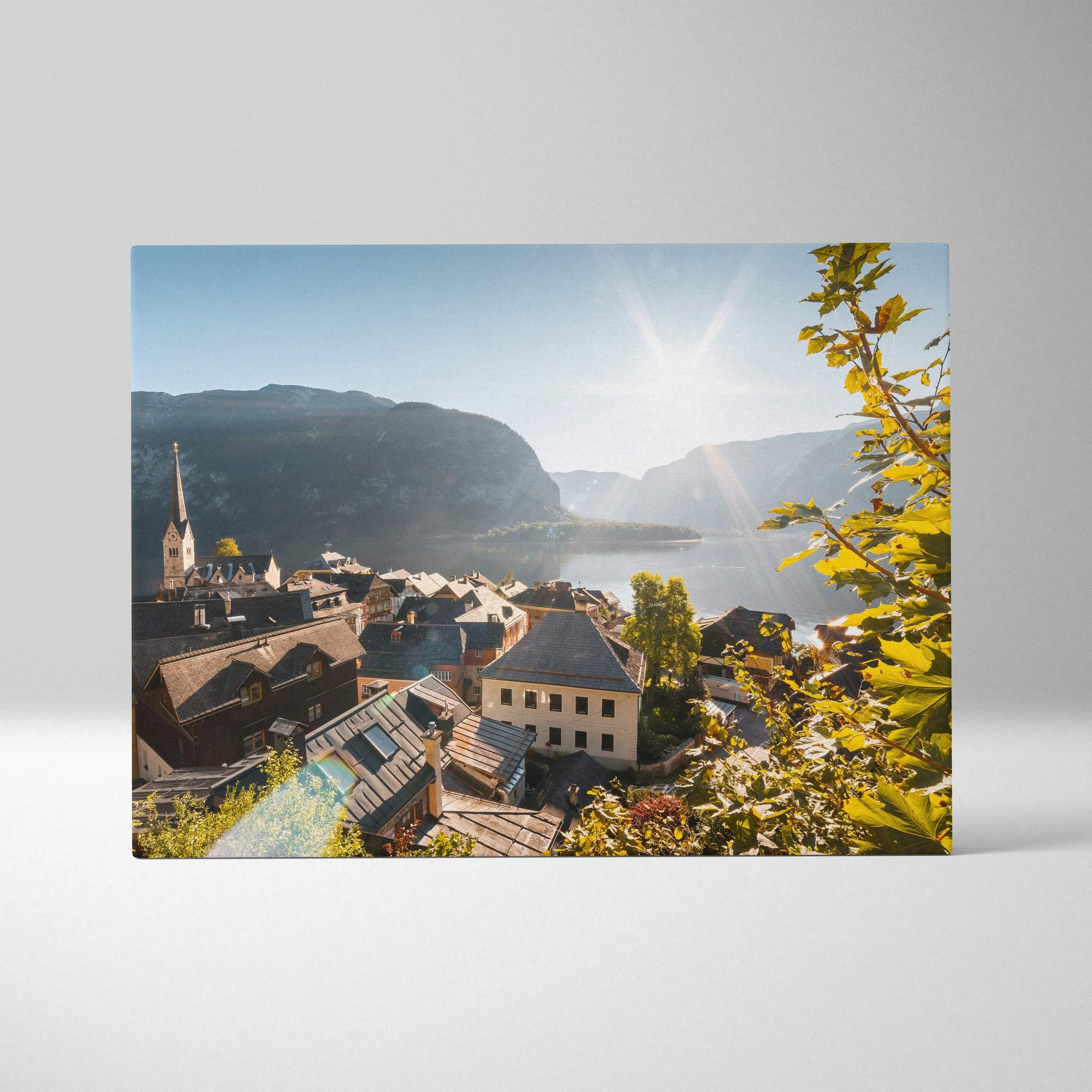 Canvas wall art featuring a sunlit village with traditional buildings, mountains, and a lake in soft natural tones.