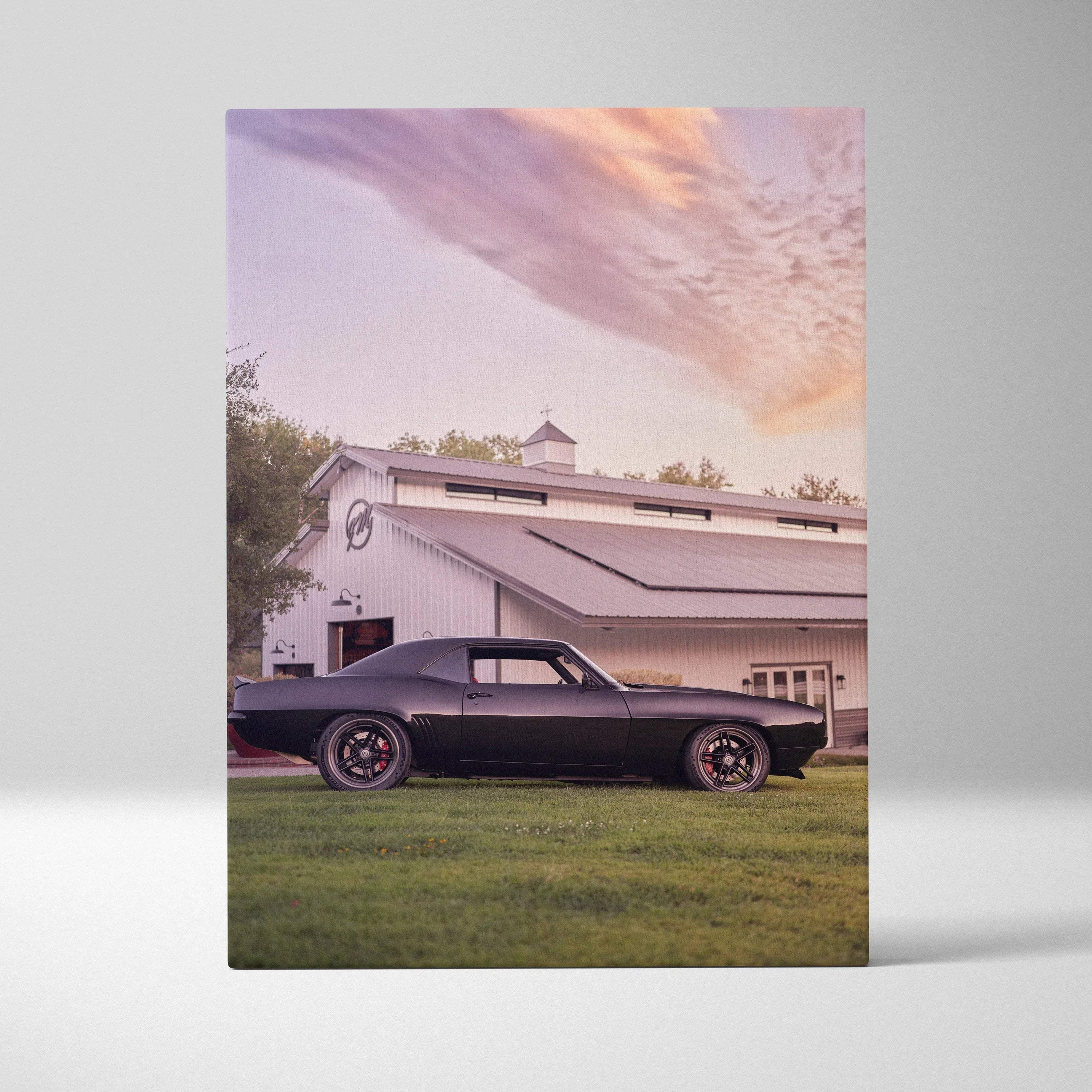 Canvas wall art featuring a sleek black 1969 camaro with red-accented wheels parked near a white barn at sunset