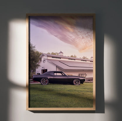 1969 black Camaro parked on grass in front of white barn under colorful sunset sky wall art poster.