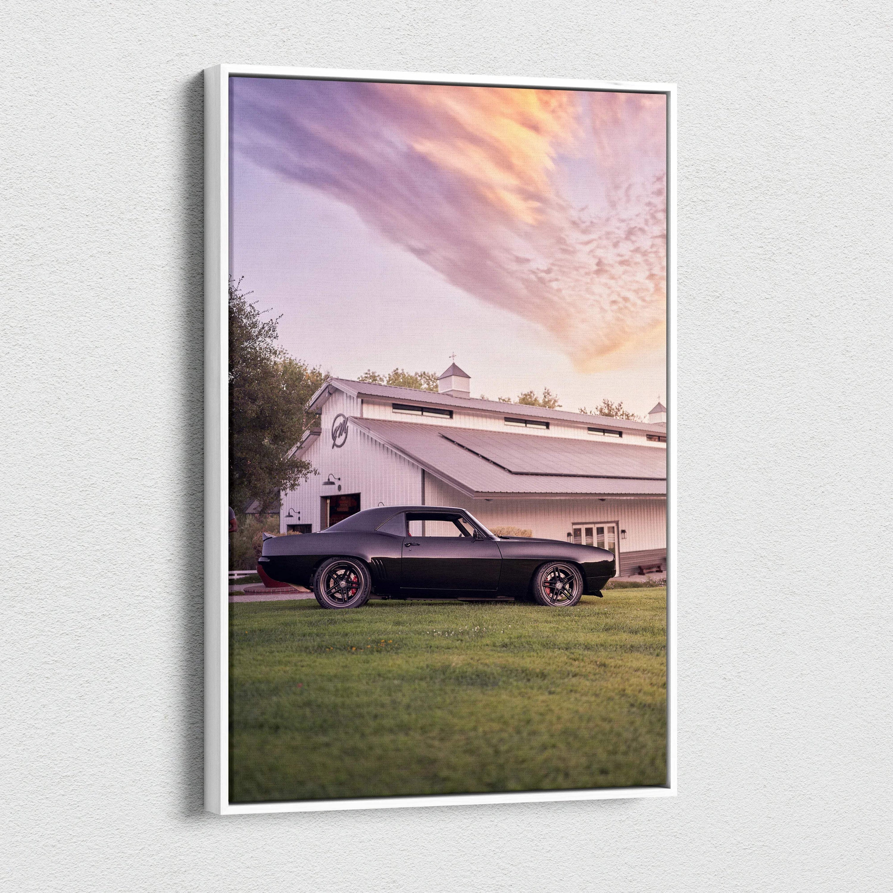 Canvas wall art featuring a sleek black 1969 camaro classic car with chrome accents parked by a white barn at sunset