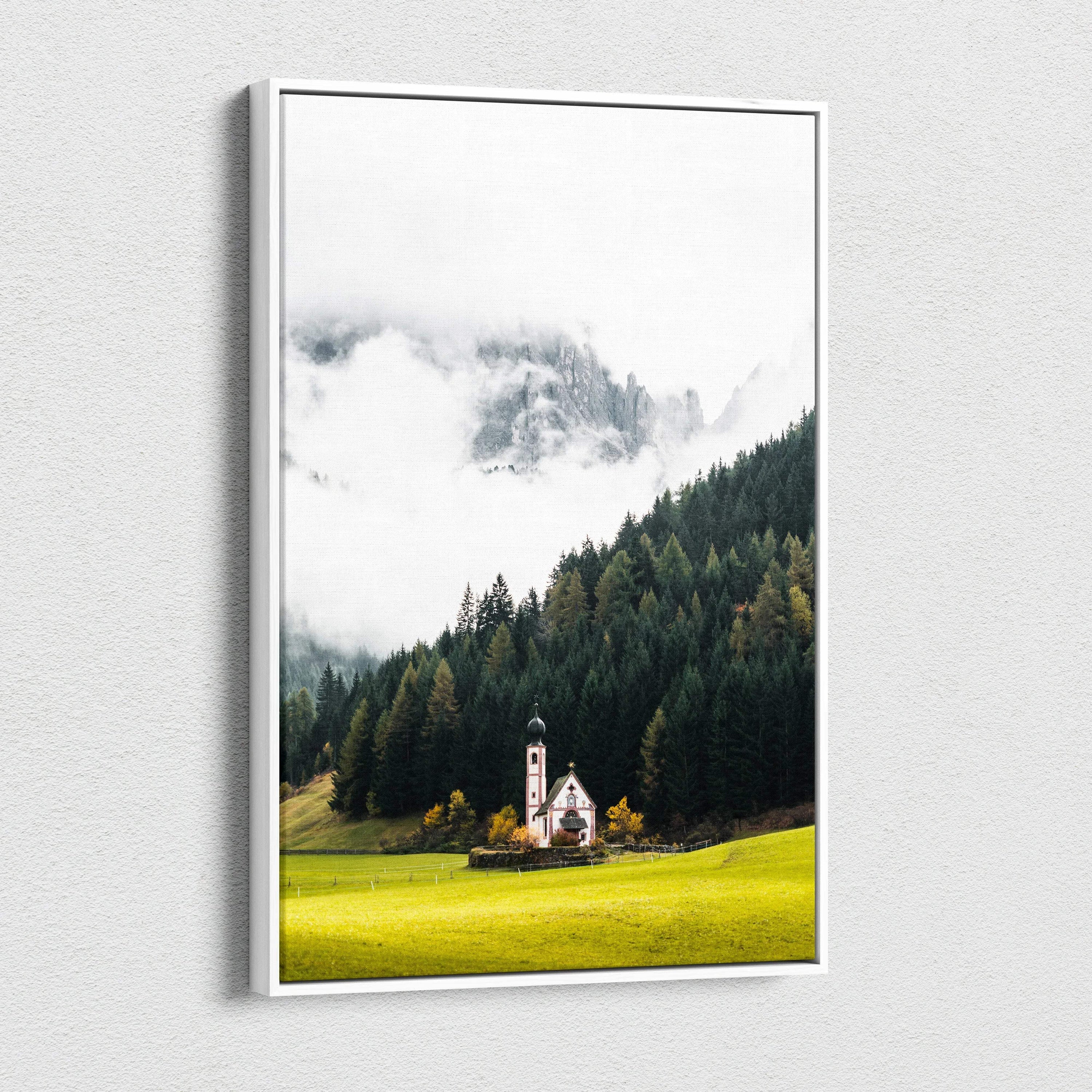 Canvas wall art featuring a small white church with a steeple surrounded by green forest and misty mountains in the.