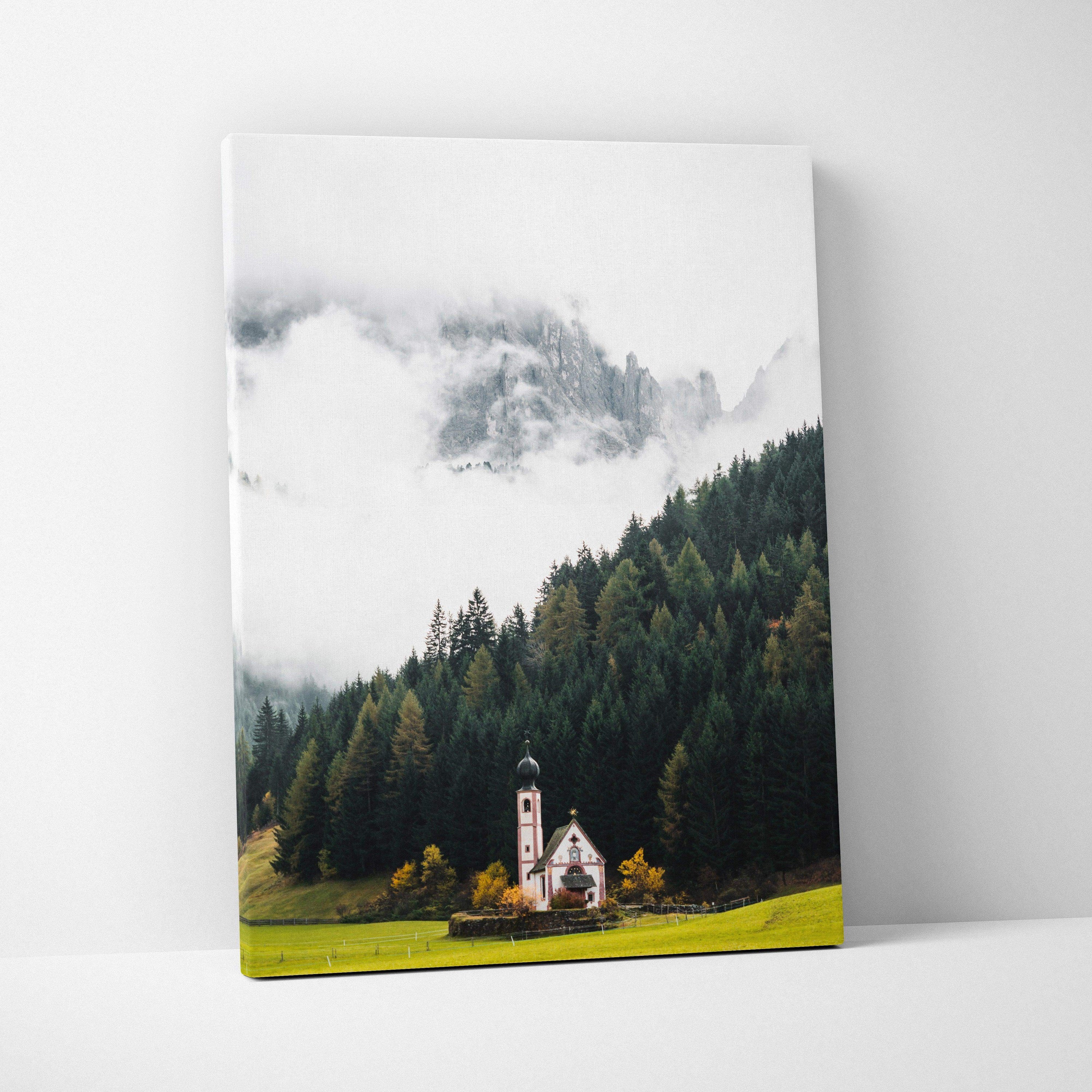Canvas art print featuring a misty mountain landscape with dense pine forest and a small church near a green meadow.