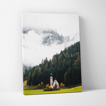 Canvas art print featuring a misty mountain landscape with dense pine forest and a small church near a green meadow.