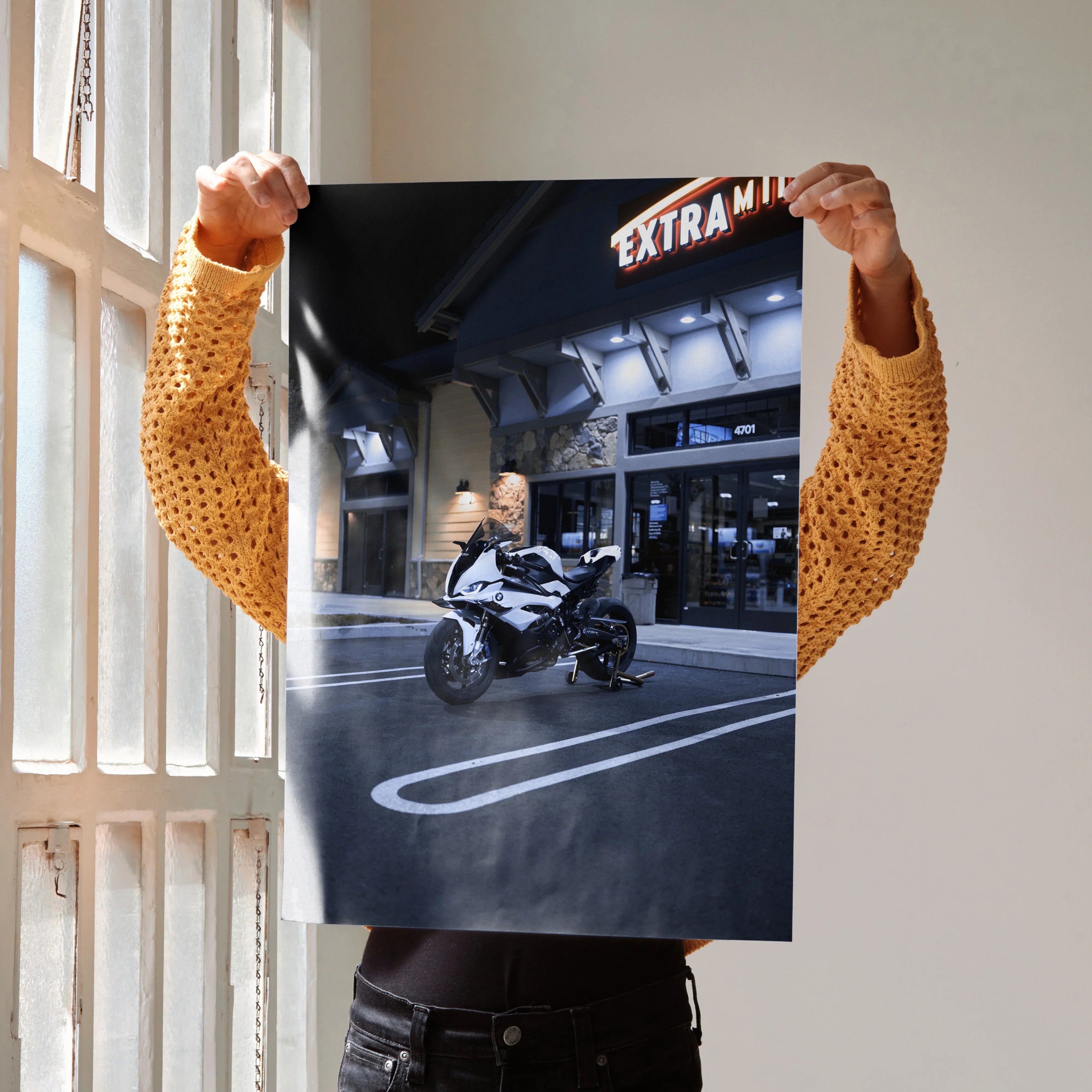 Black and white BMW S1000RR motorcycle poster held indoors, nighttime urban wall art with neon sign backdrop.