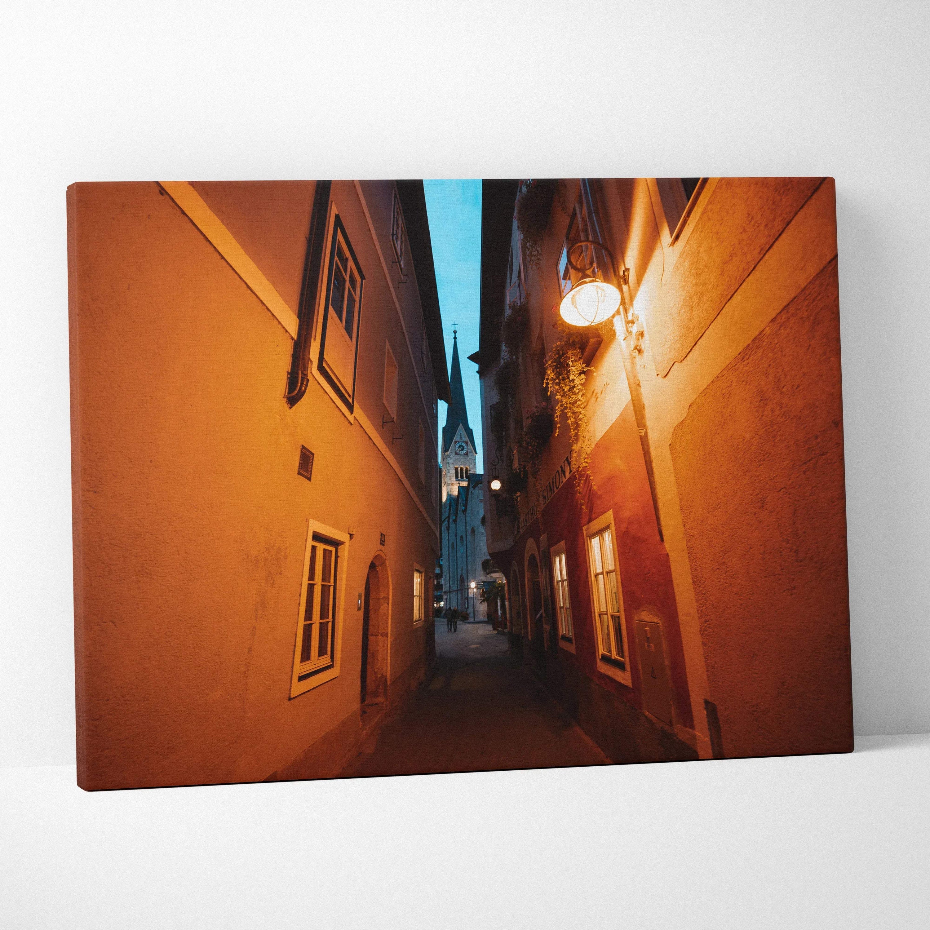 Canvas wall art featuring a narrow alley between orange buildings under a blue evening sky with warm street lighting.
