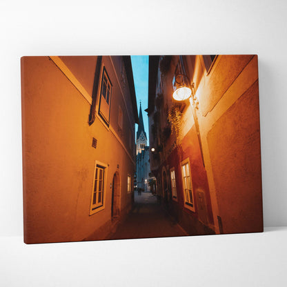 Canvas wall art featuring a narrow alley between orange buildings under a blue evening sky with warm street lighting.