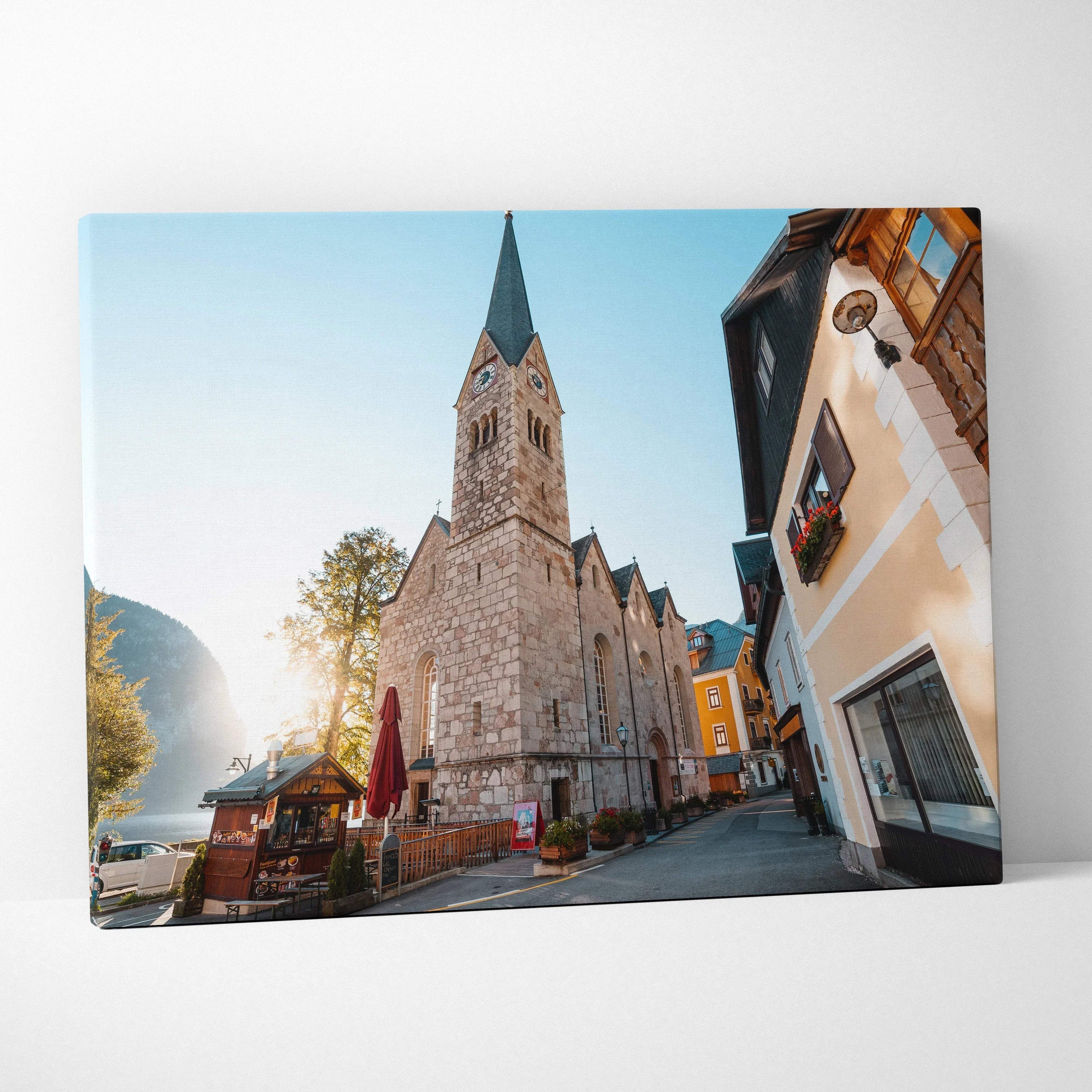 Canvas wall art featuring a tall stone church with a pointed steeple, warm sunlight, and quaint European street buildings.