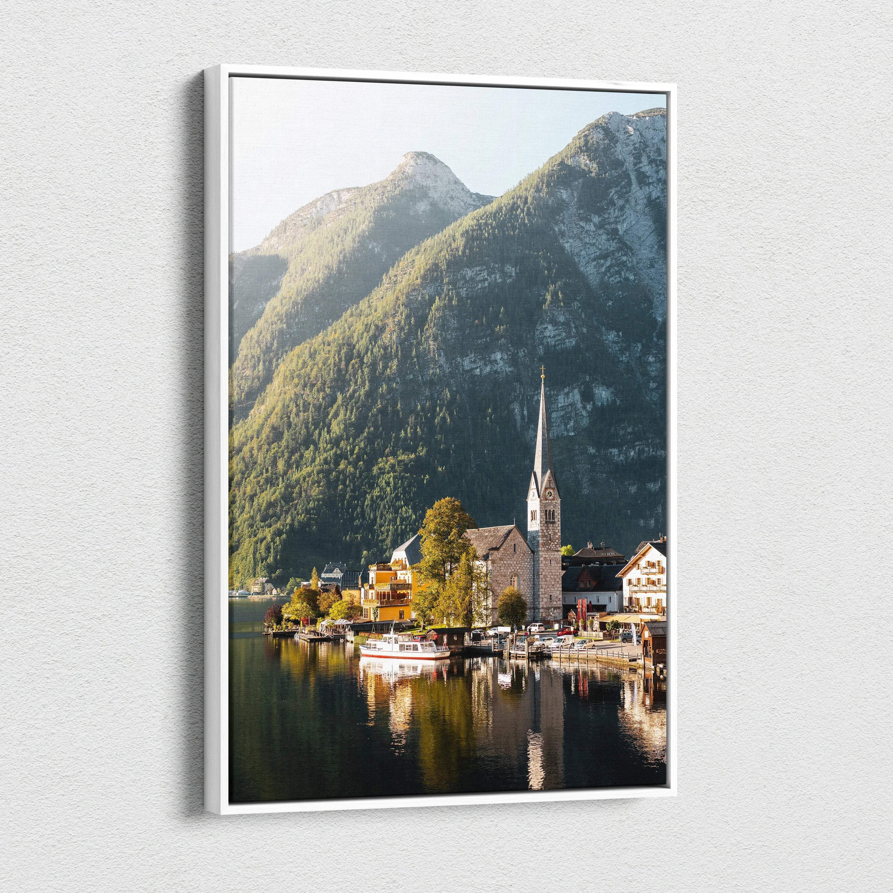 Framed canvas wall art showing a serene lakeside village with a church steeple, surrounded by forested mountains and.
