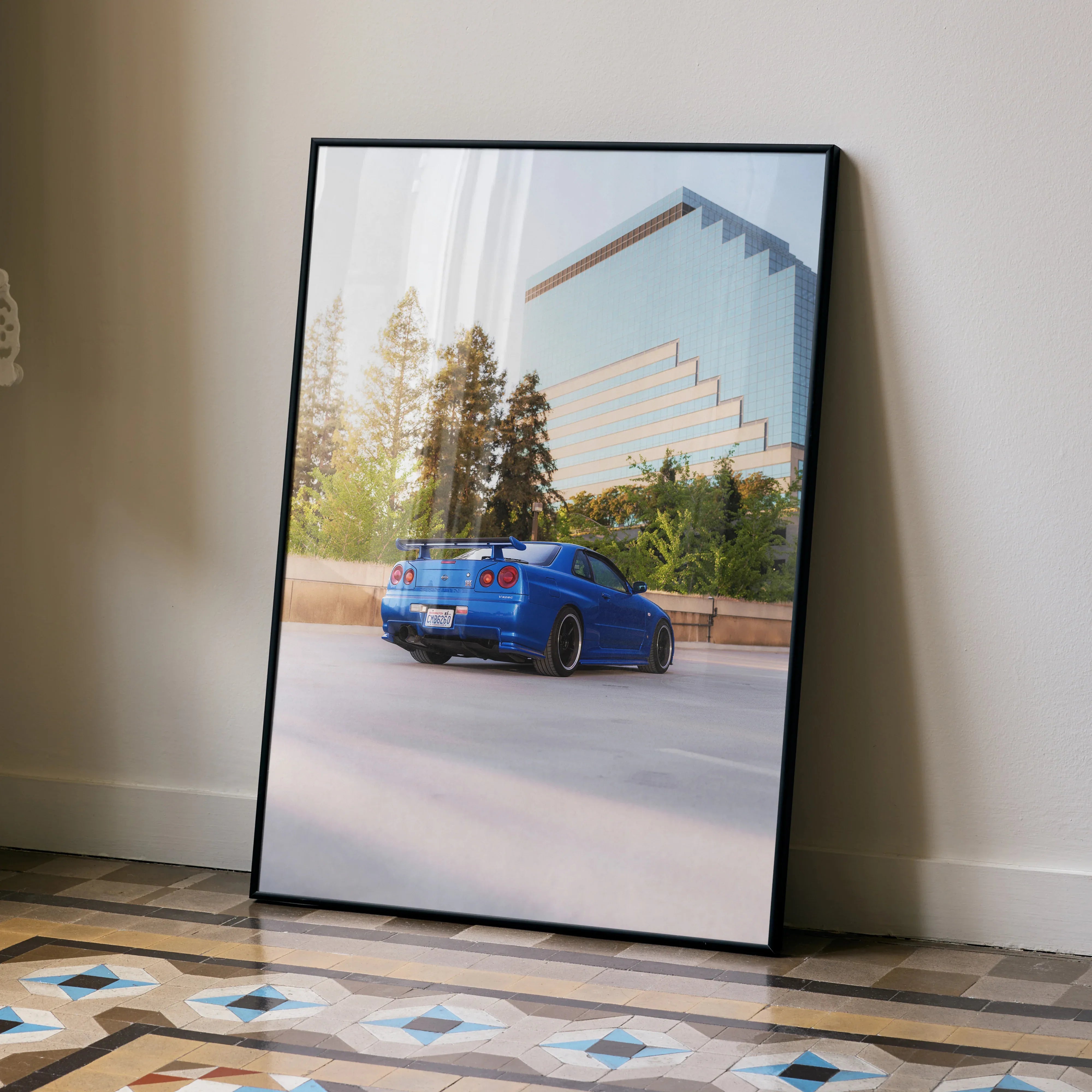 Nissan R34 GTR Skyline blue car poster leaning against beige wall, modern building and trees background wall art.