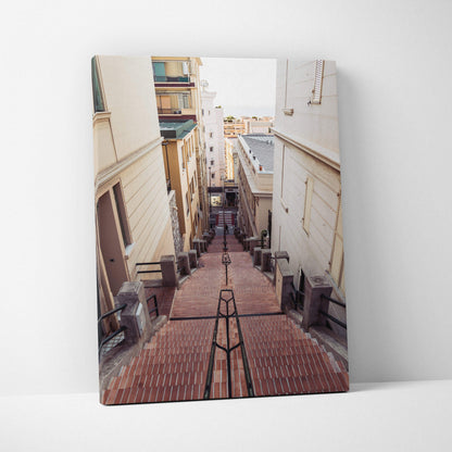 Vertical canvas wall art featuring a narrow urban staircase between beige buildings with red brick steps and metal handrails.