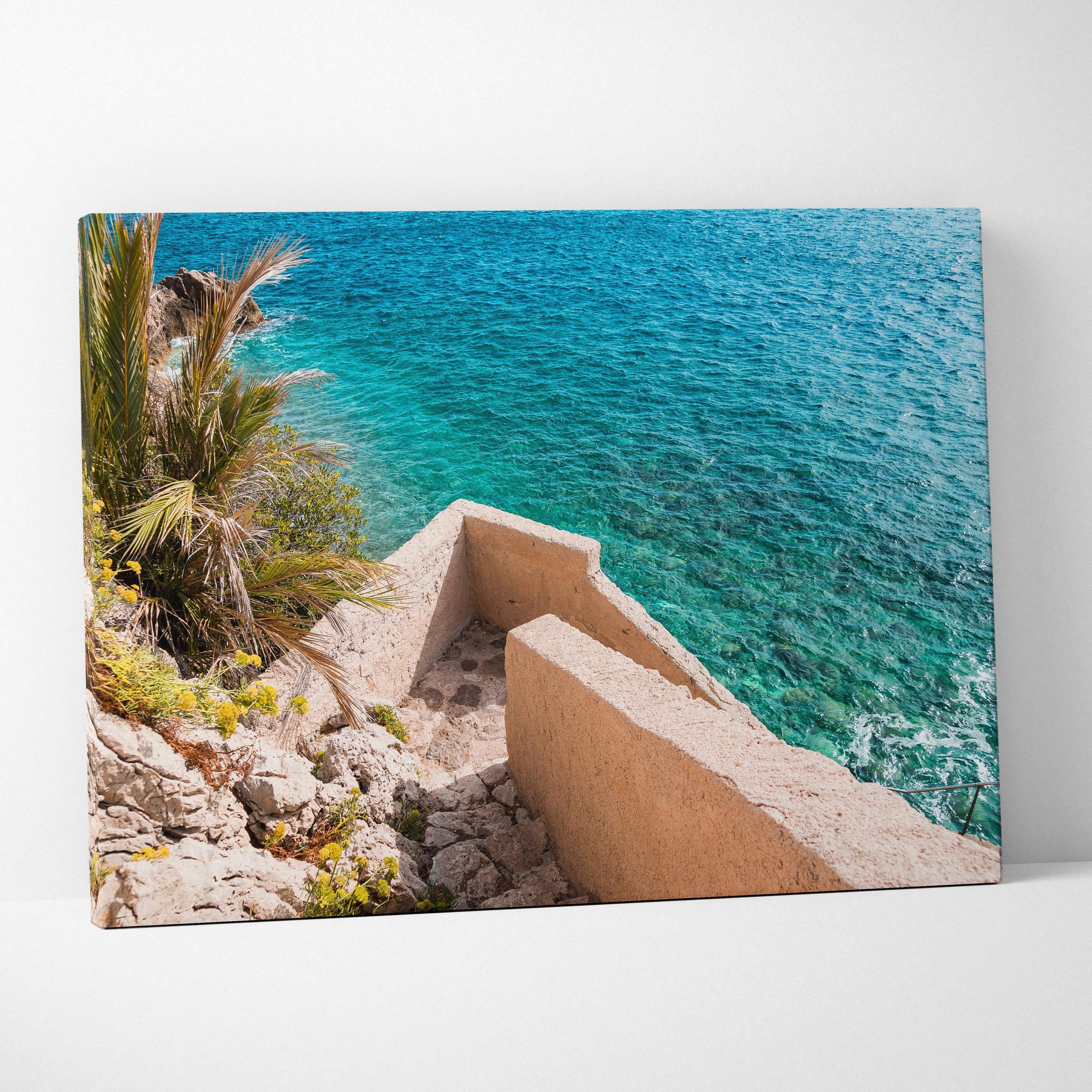 Canvas wall art featuring a coastal scene with beige stone architecture and vibrant blue ocean water.