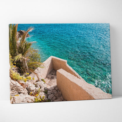 Canvas wall art featuring a coastal scene with beige stone architecture and vibrant blue ocean water.