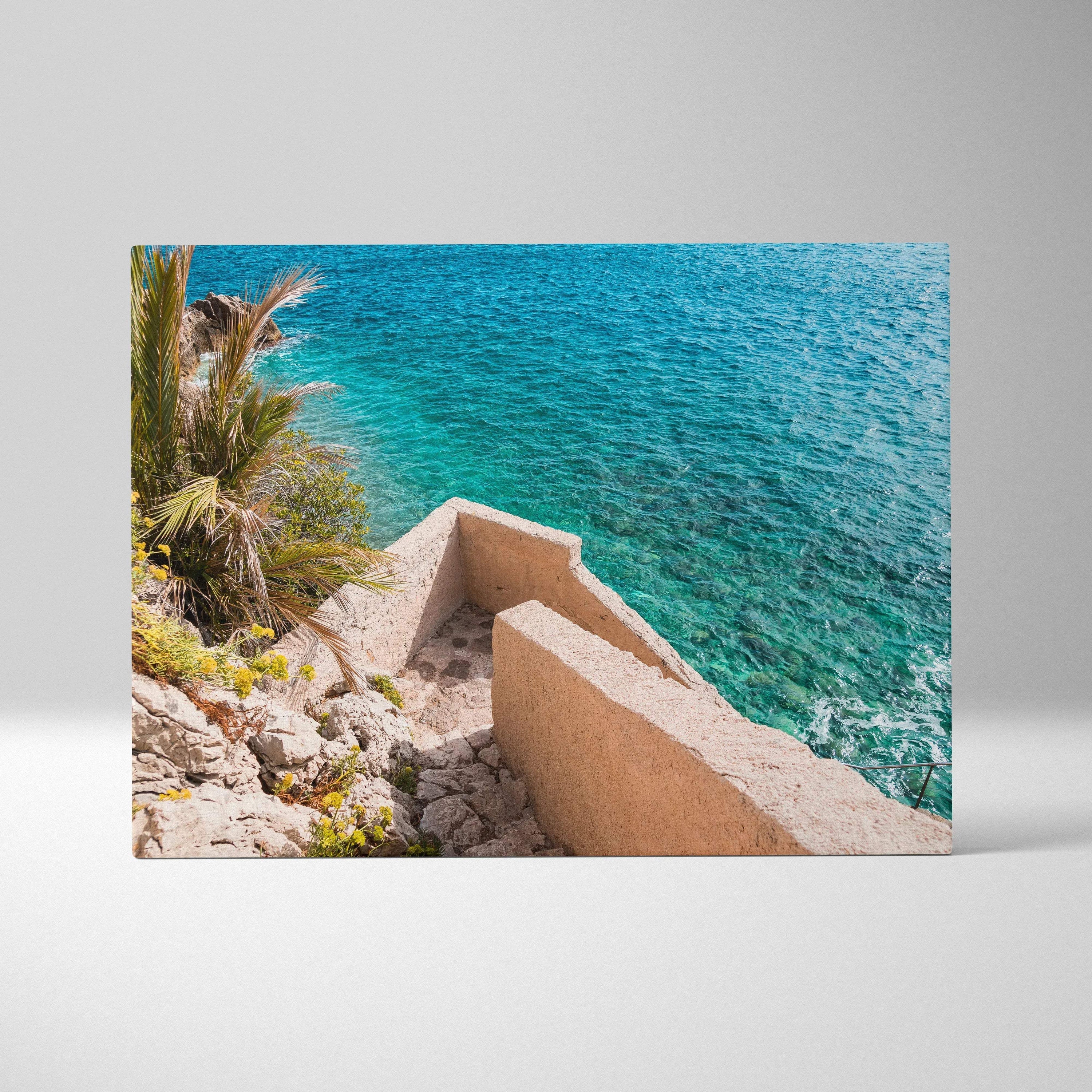 Canvas wall art featuring a coastal scene with turquoise water, rocky shore, palm tree, and beige concrete stairs.