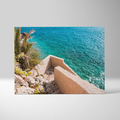 Canvas wall art featuring a coastal scene with turquoise water, rocky shore, palm tree, and beige concrete stairs.