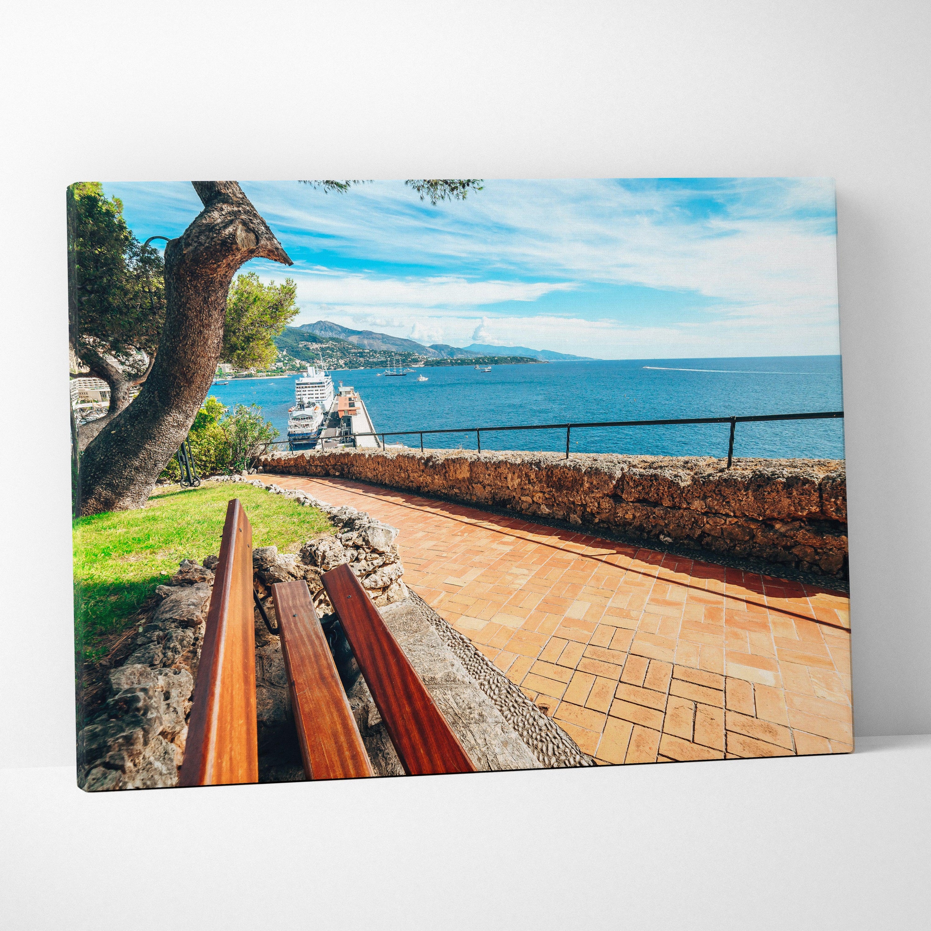 Canvas wall art of a sunny beachside scene with a wooden bench, stone path, and blue ocean under a partly cloudy sky.