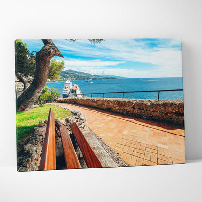 Canvas wall art of a sunny beachside scene with a wooden bench, stone path, and blue ocean under a partly cloudy sky.