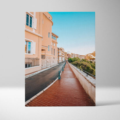 Vertical canvas wall art of a sunlit street lined with peach buildings and a clear blue sky.