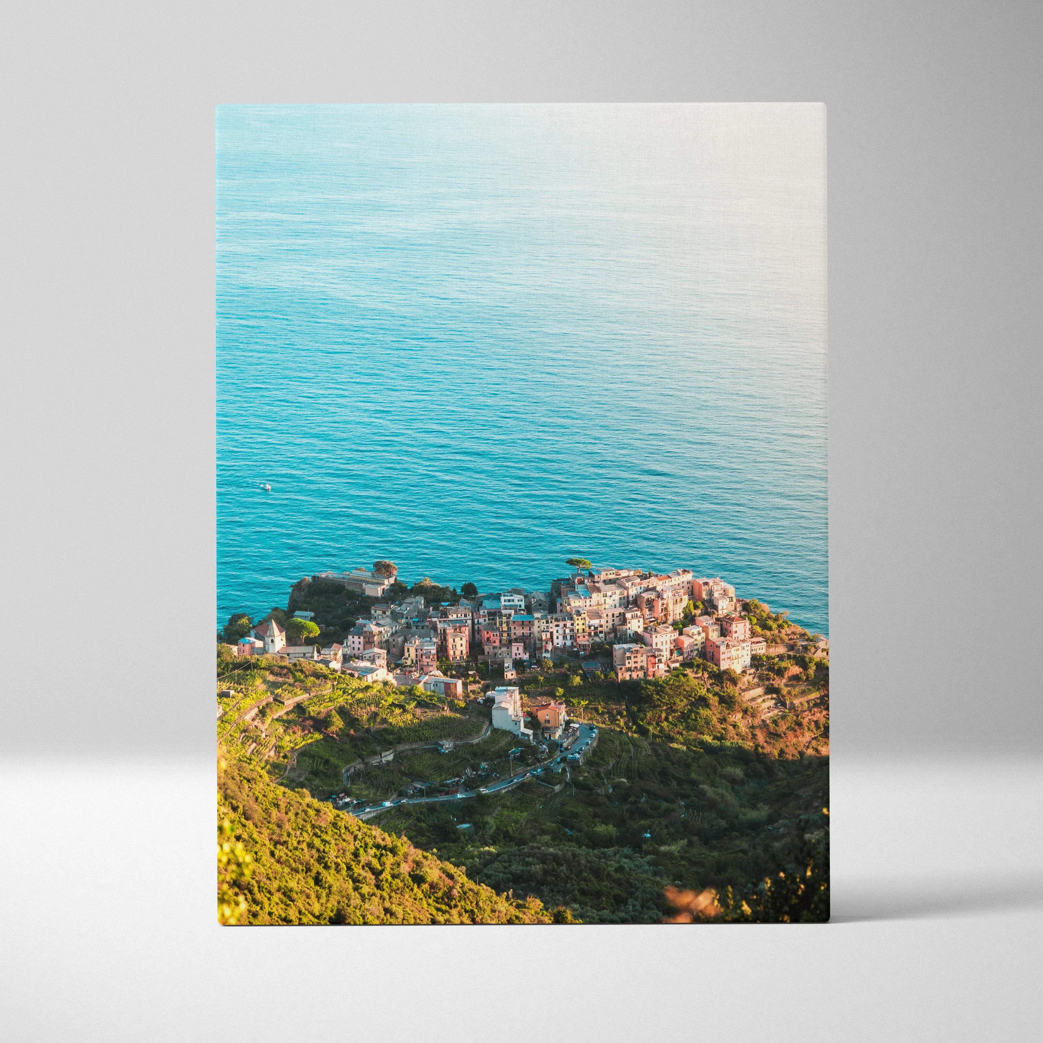 A rectangular canvas print of coastal architecture with terracotta roofs on green hills by turquoise ocean waters