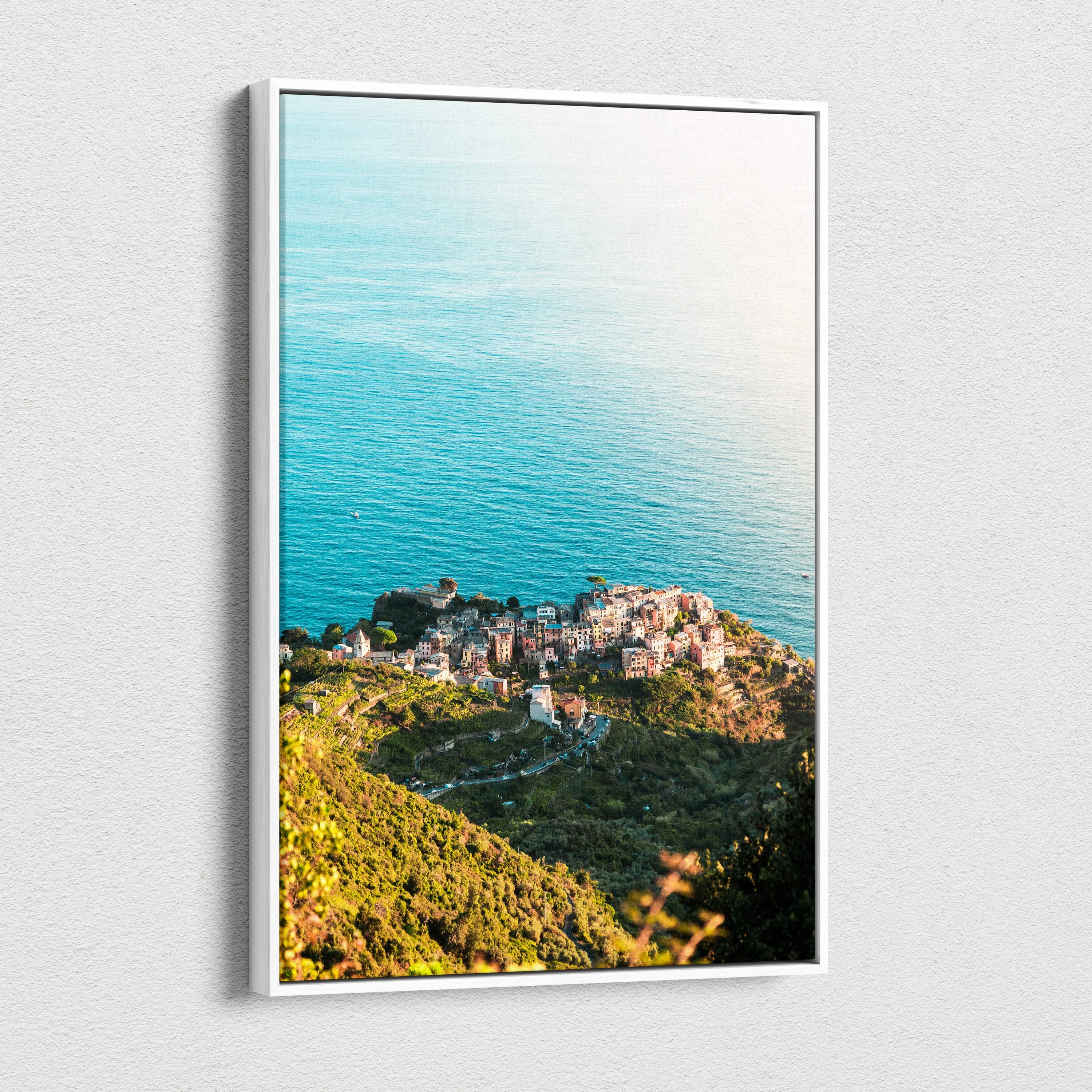 Framed canvas wall art featuring sunlit coastal town with green hills and calm blue ocean background.
